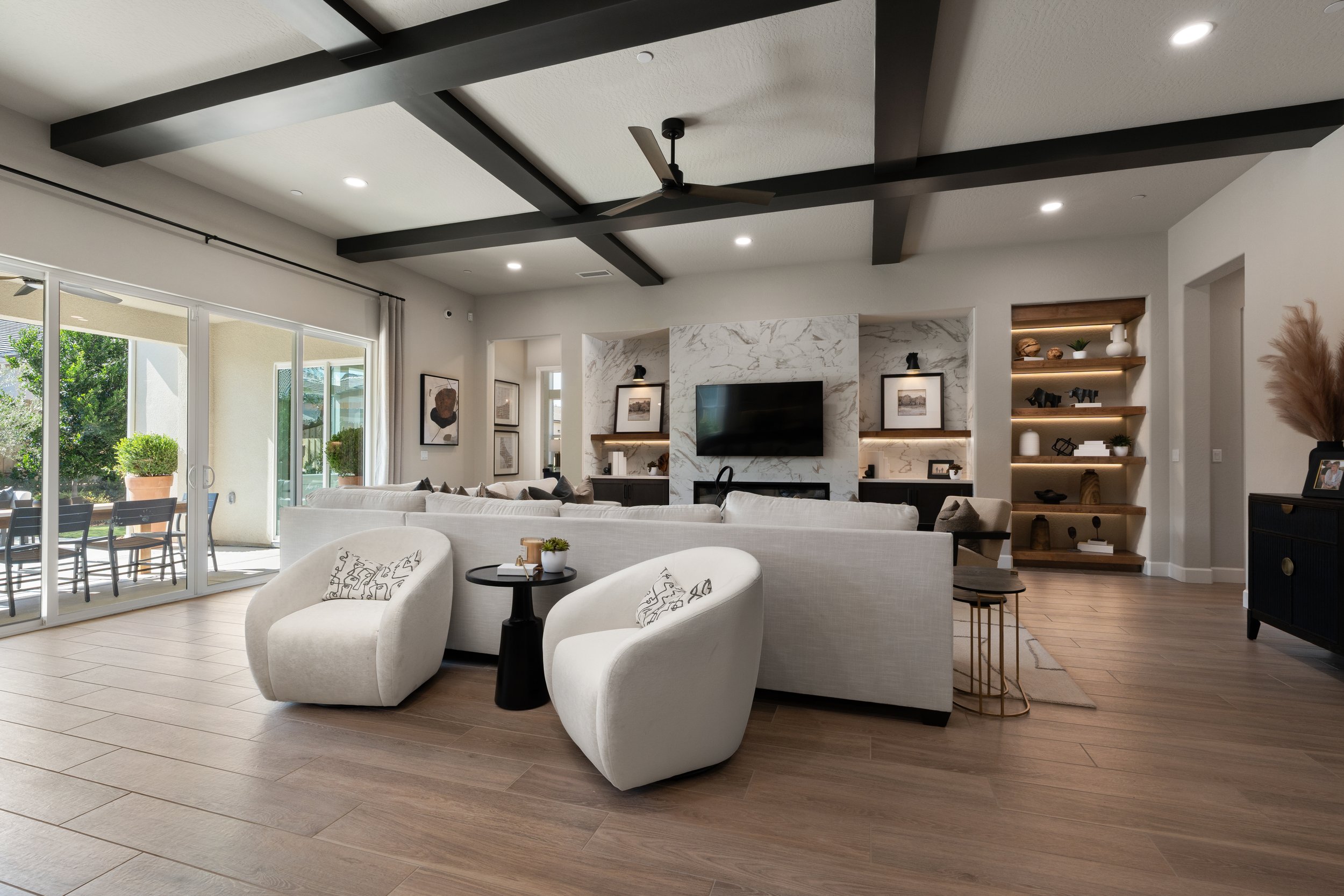 Modern living room with white sofa, armchairs, and a black side table, large sliding glass doors leading to a patio with outdoor dining, black ceiling fan, and built-in shelves with decor, artwork, and lighting.