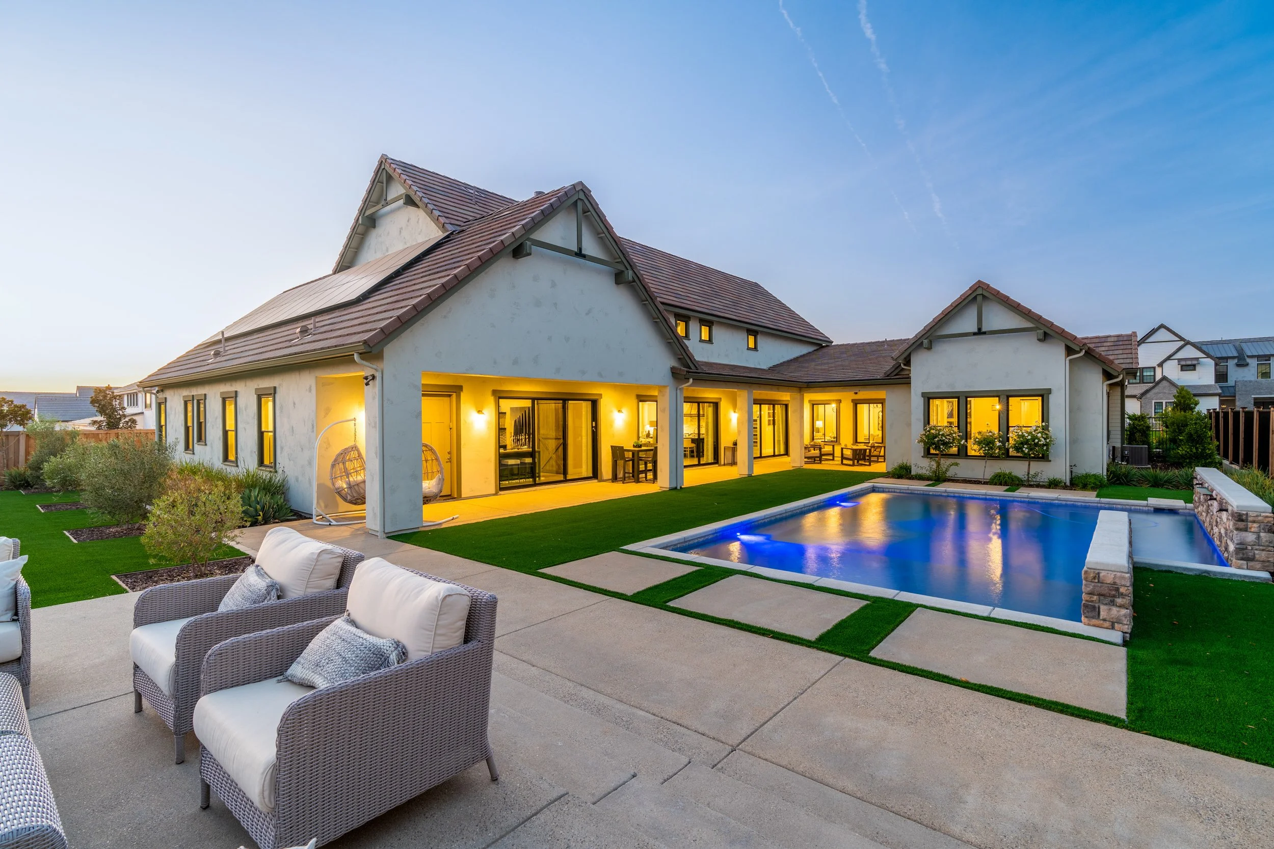 View of a modern house with illuminated interior, a backyard swimming pool, outdoor seating area with wicker furniture, and a landscaped garden during dusk.