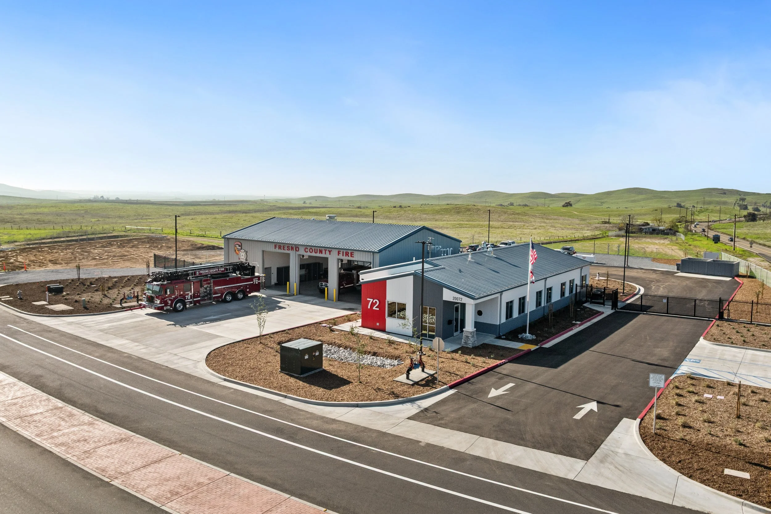 A fire station with a fire truck parked outside, surrounded by a parking lot and landscaped areas, with green rolling hills and a blue sky in the background.