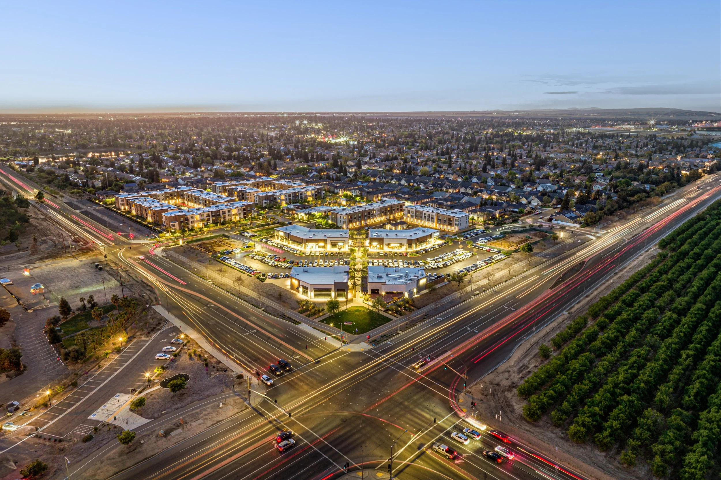 Aerial view of a city at dusk showing illuminated buildings, streets with light trails from moving cars, residential neighborhoods, and surrounding hills in the distance.