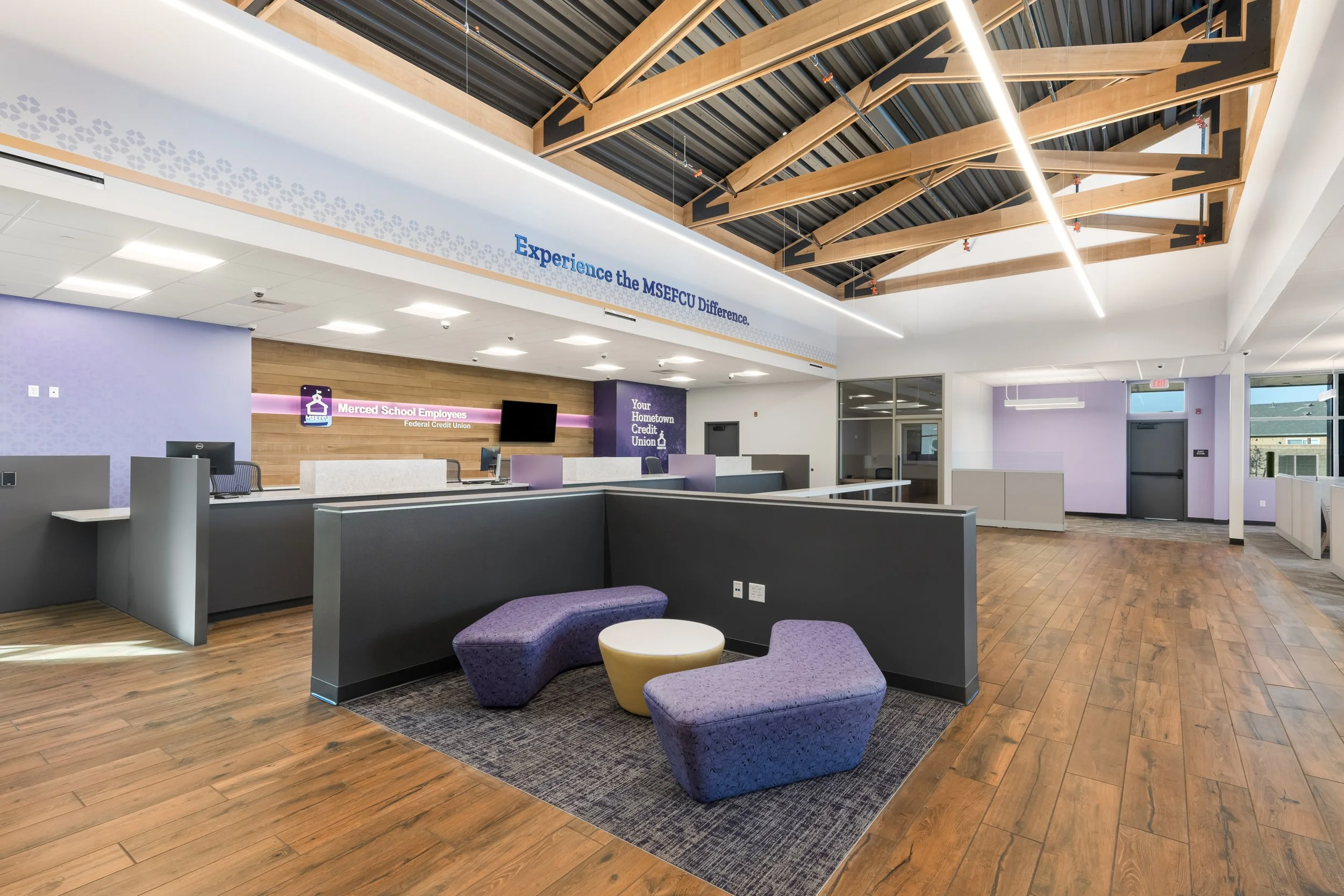 Interior of a modern financial institution with a reception area, purple and grey furniture, hardwood flooring, and exposed wooden ceiling beams, featuring signs for Merced School Employees Federal Credit Union and information about the institution.