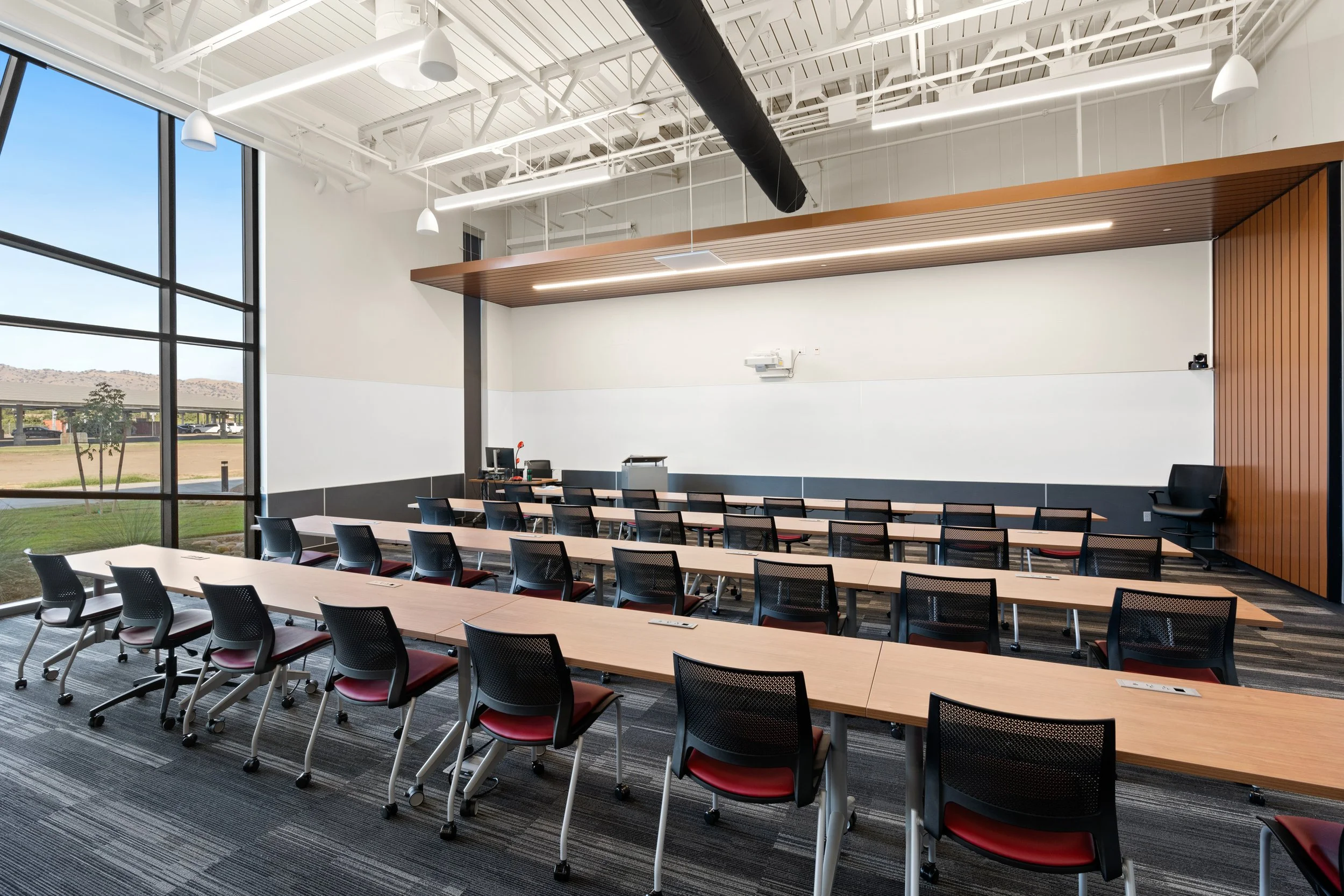 Interior of a modern conference room with multiple rows of tables and chairs, large windows showing an outside landscape, and ceiling with exposed rafters, pipes, and lighting.