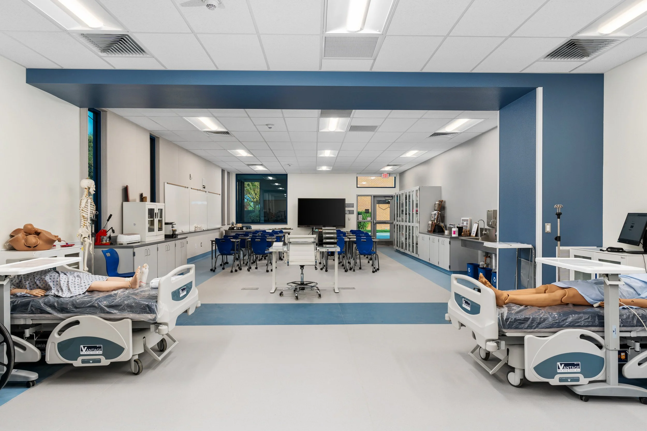 Medical training classroom with two hospital beds, medical mannequins, and medical equipment, with chairs and a large screen at the front.