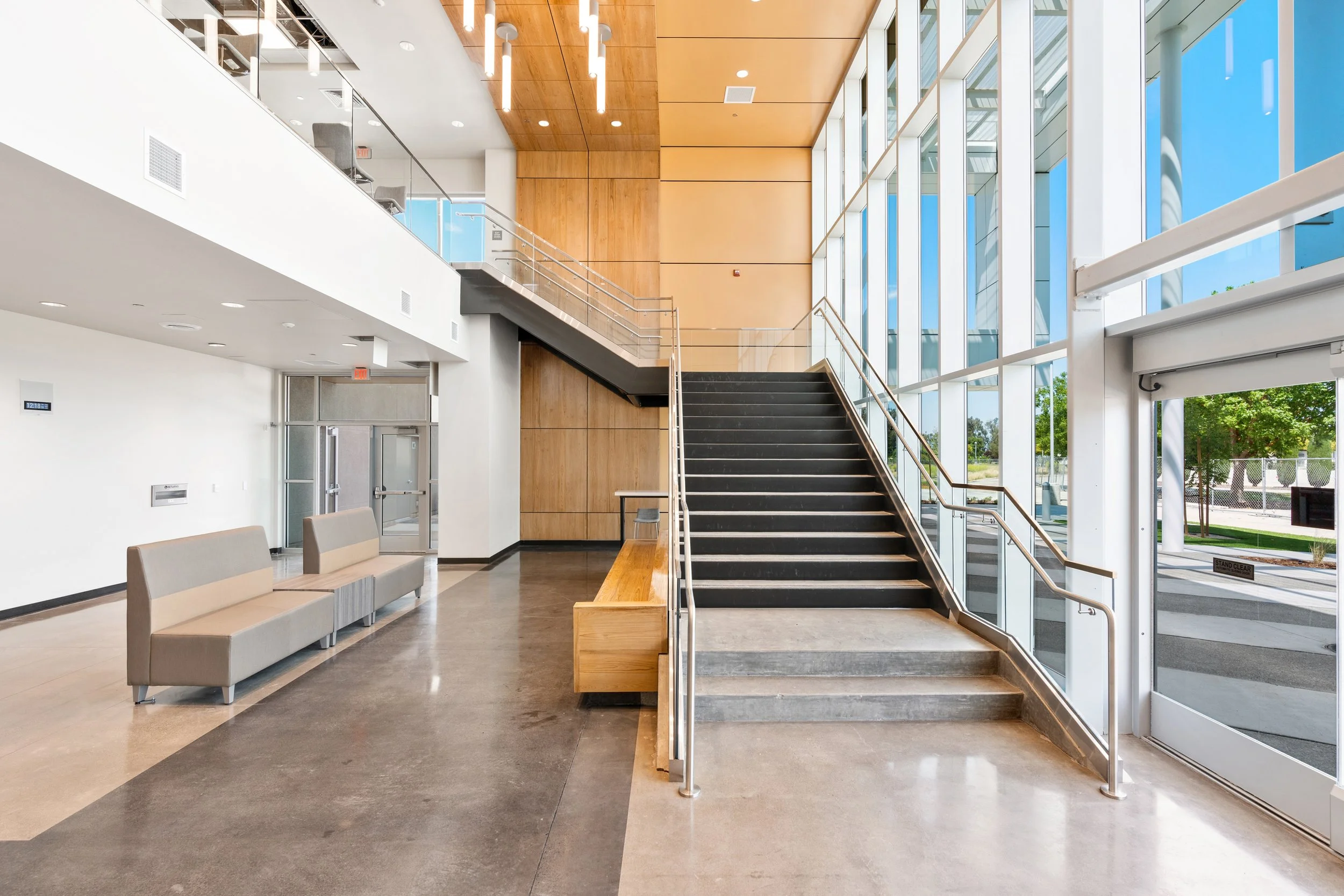 Bright modern lobby with large glass windows, a staircase, seating area with beige couches, and wooden wall panels.