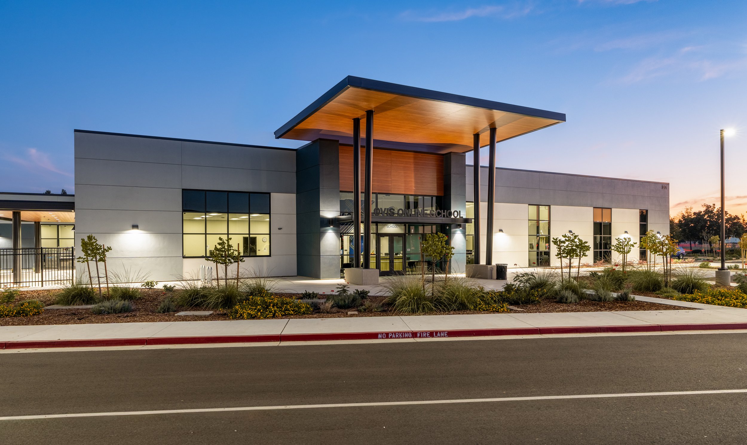 Modern school building with large windows and a prominent entrance. The building has a flat roof with an overhanging wooden canopy supported by tall black columns. There are small trees and shrubs in front, with a sidewalk and a street marked with a 