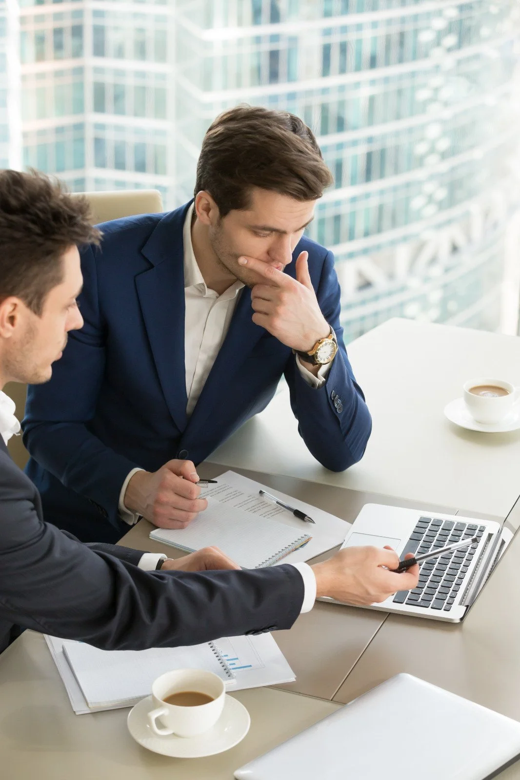 Two businessmen in suits having a serious discussion at a conference table with laptops, documents, and coffee cups in a modern office with large windows.