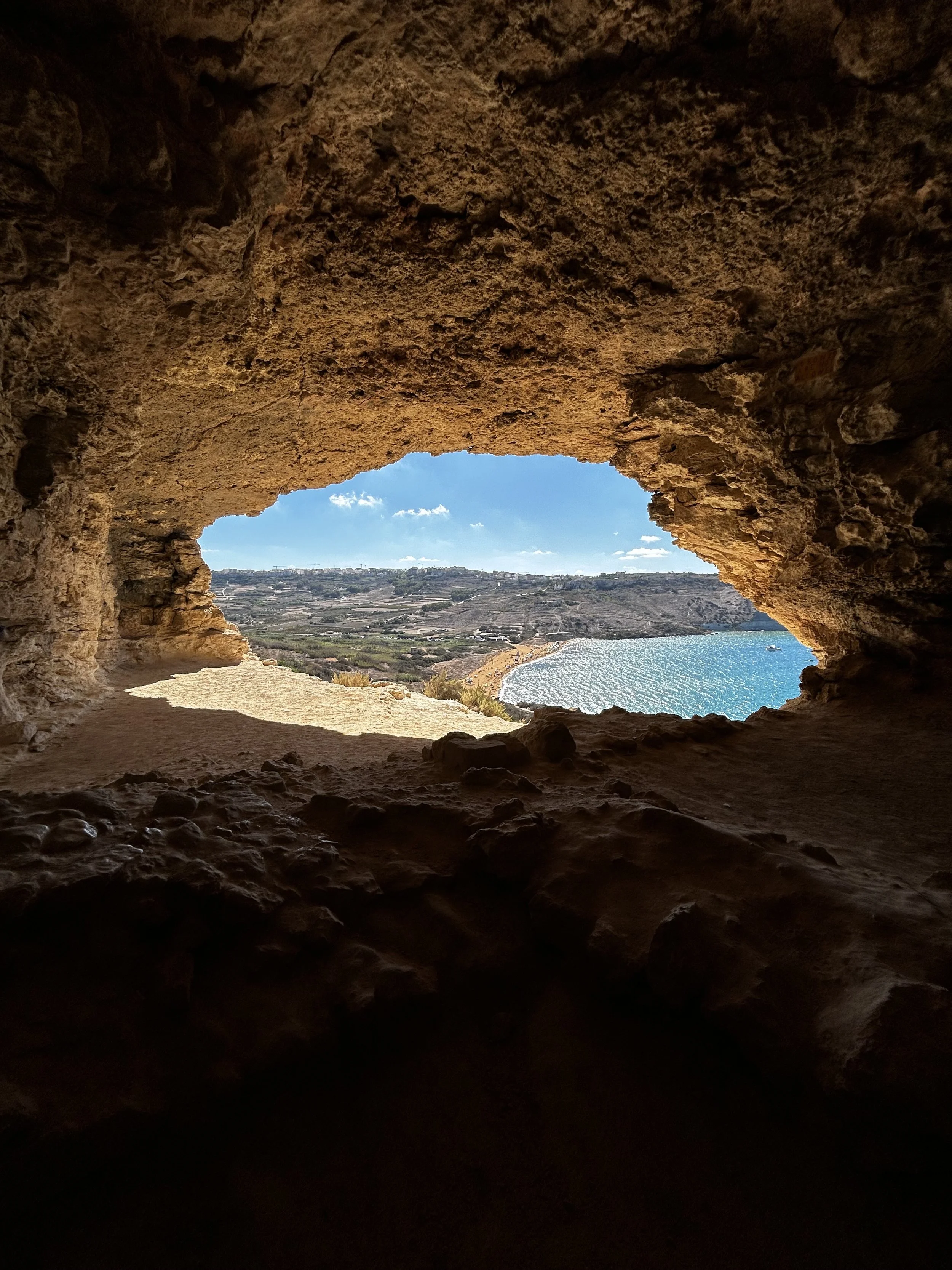 View through a cave opening showing a scenic coastline with water, shoreline, and distant land under a partly cloudy sky.
