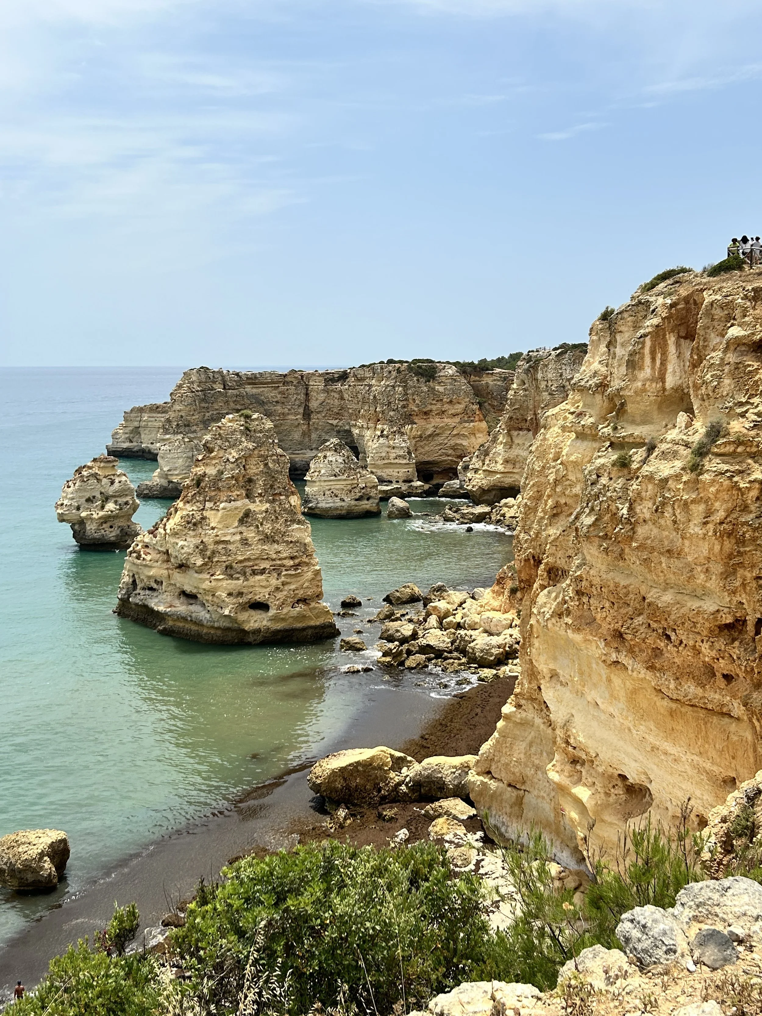 Cliffs along the coastline with large rock formations in the water, greenery at the bottom, and people standing on a viewing platform on top of the cliffs.