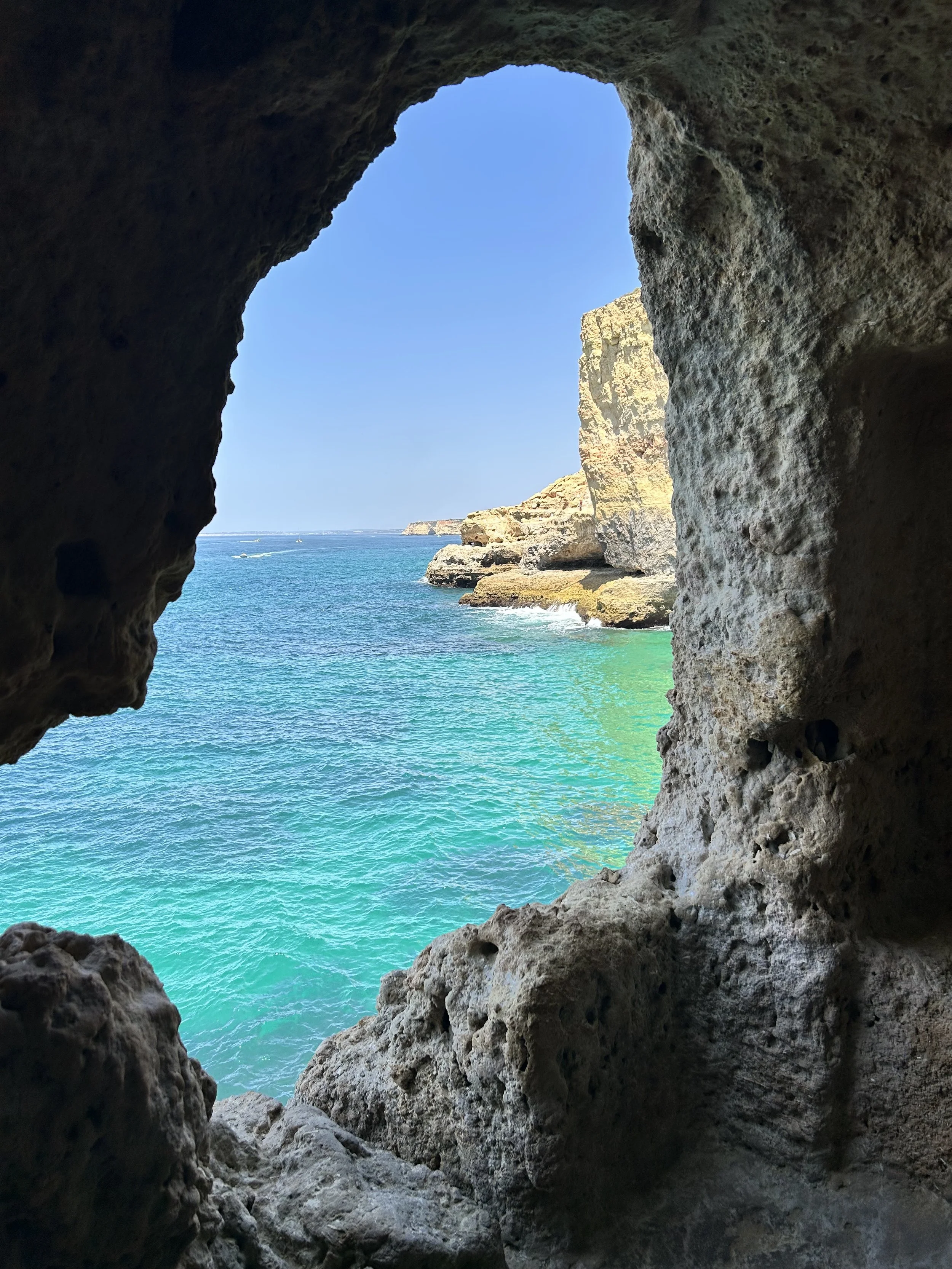 View of blue ocean water through a natural rock window with cliffs and small boats in the distance at Algar Seco.