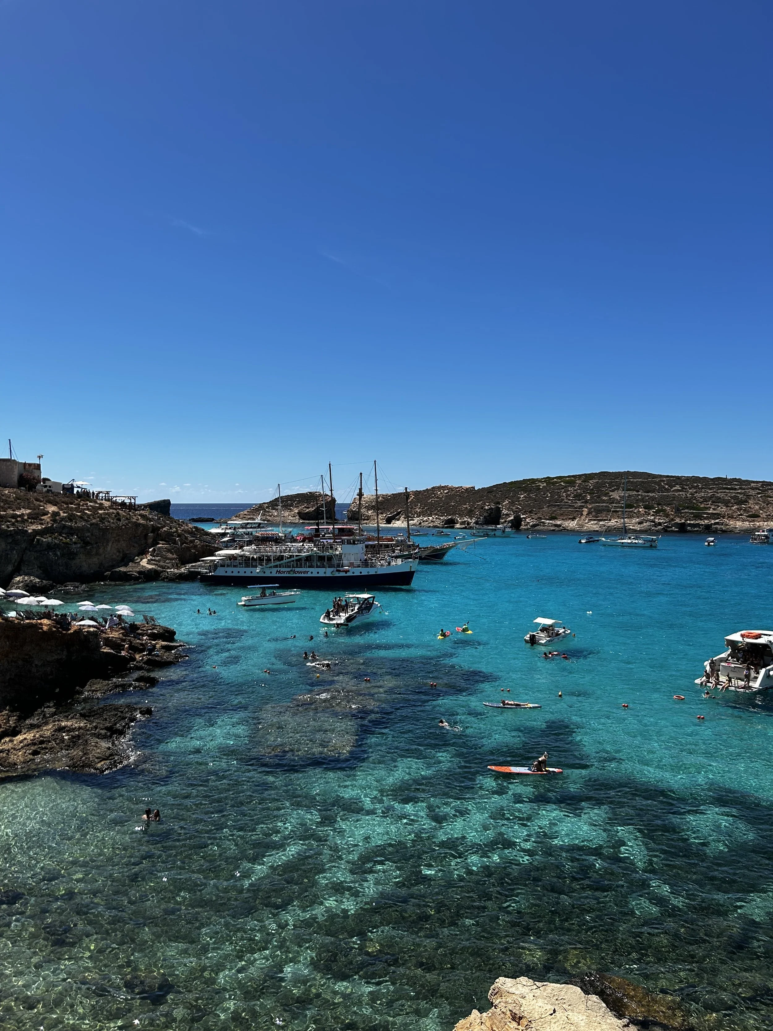 A coastal scene with turquoise water, several boats anchored, and people swimming and paddleboarding. Rocky shores and a clear blue sky in the background on Comino Island, Malta.