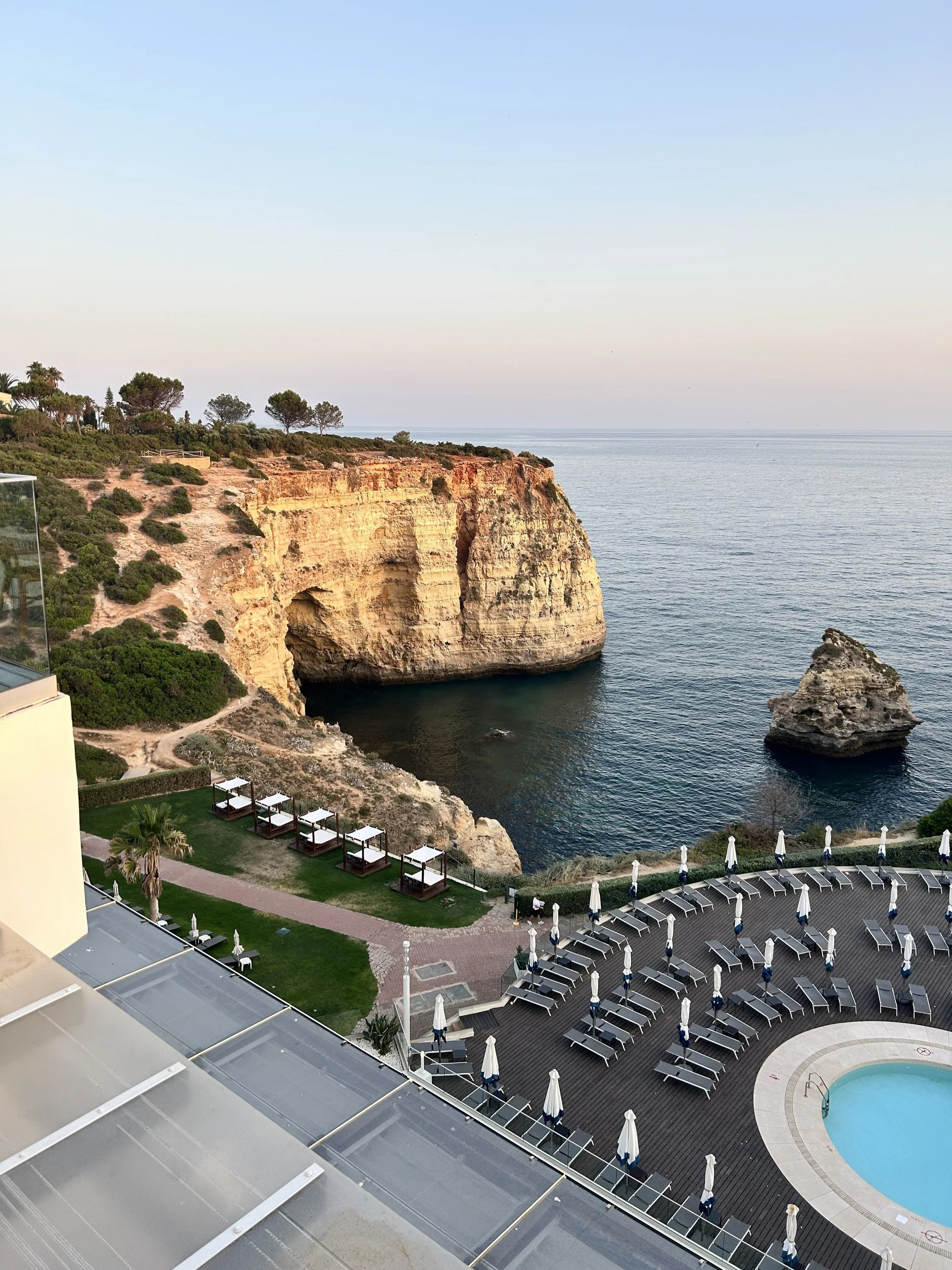 View of a seaside resort with a pool, lounge chairs, and cabanas overlooking cliffs and the ocean at sunset.