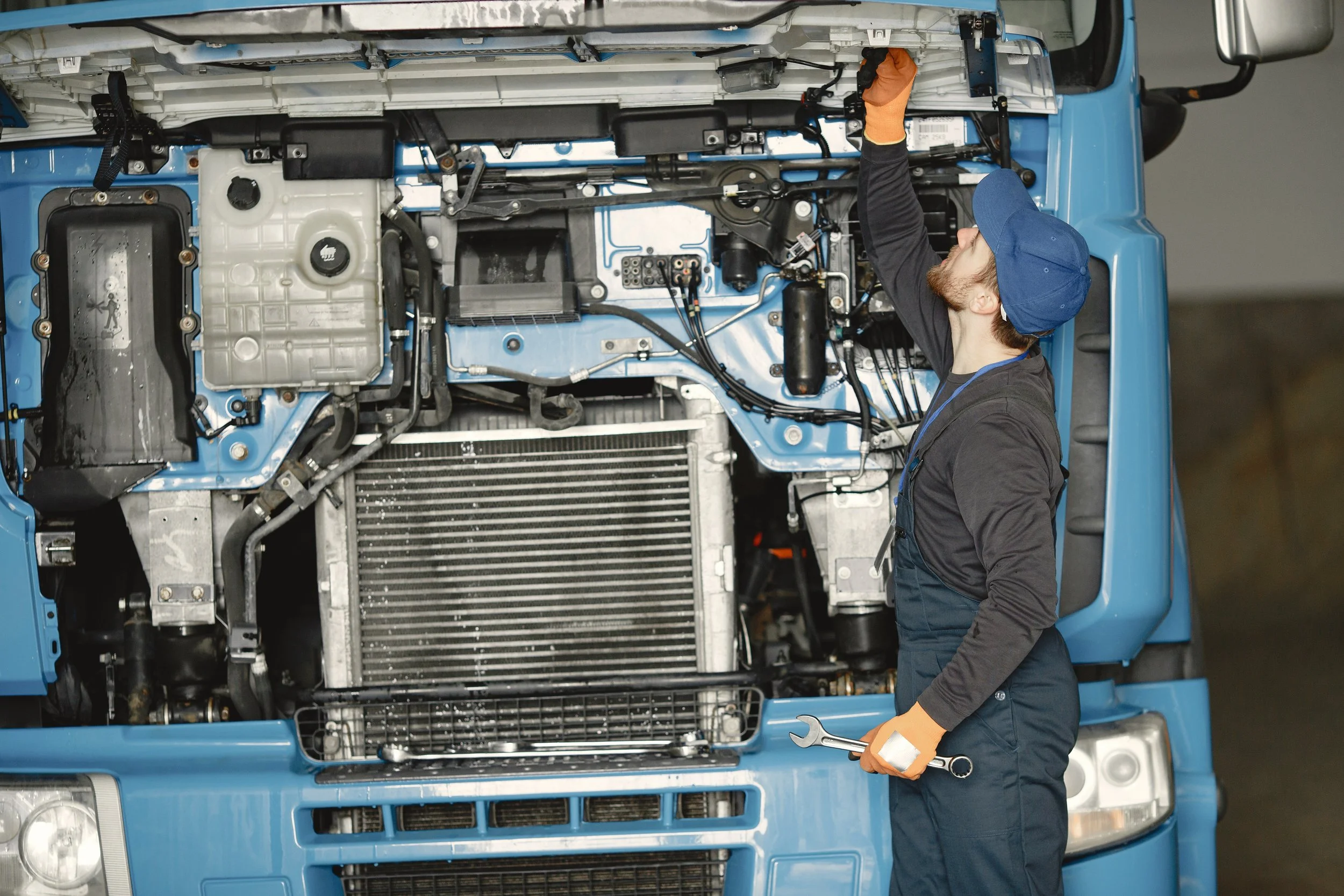 A male mechanic with a blue cap and black uniform working on the engine of a blue truck, using a wrench and inspecting the engine components.