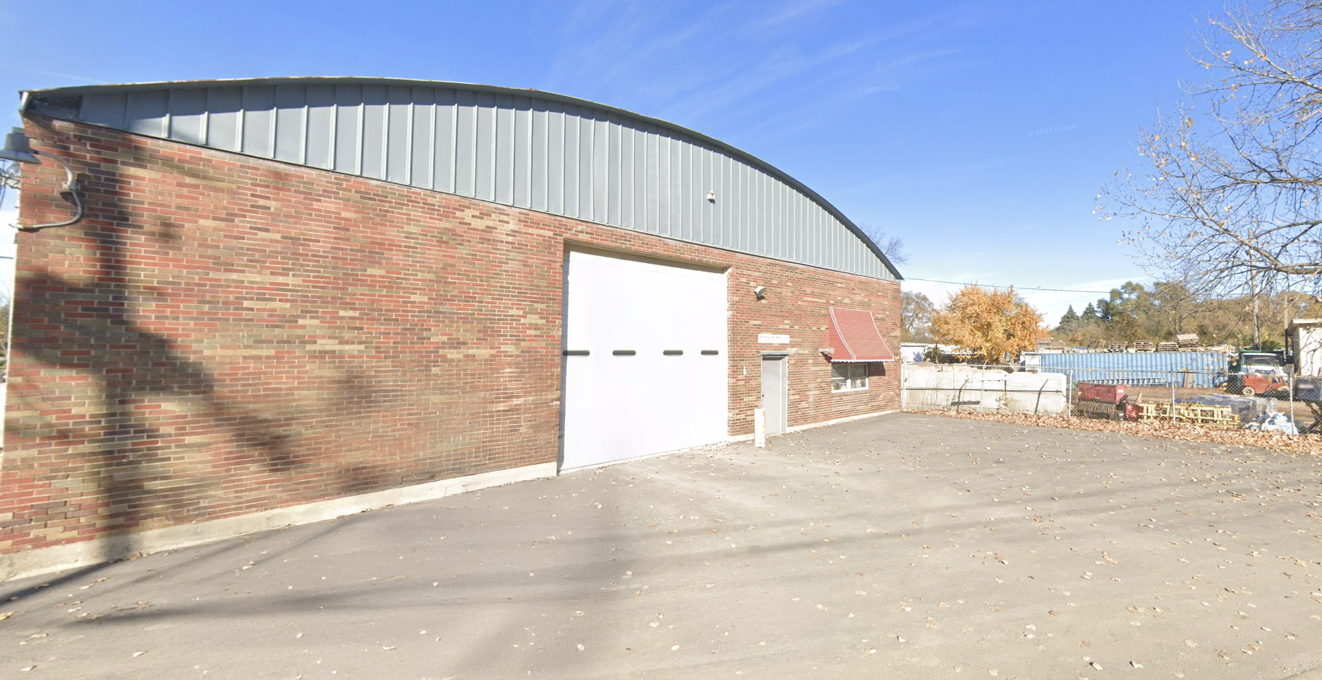 A large brick building with a metal roof, a white garage door, and a small side door with an awning. The area surrounding the building is paved, with fallen leaves and a partly cloudy blue sky. In the background, there are trees with autumn foliage and some industrial equipment.
