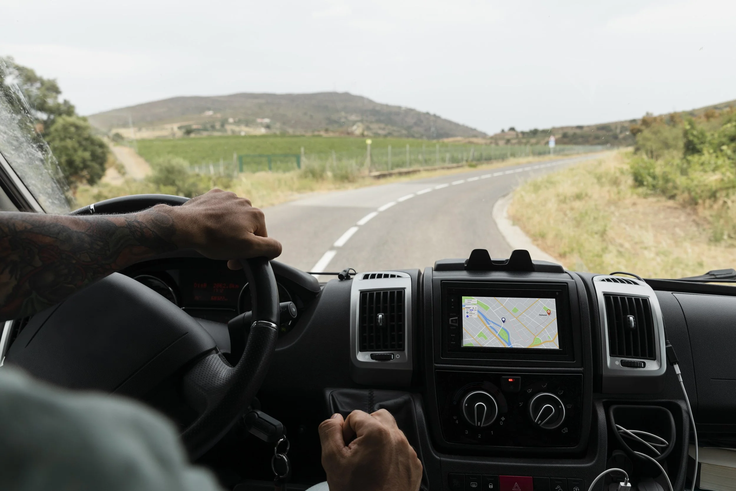 Interior of a vehicle showing a driver with tattoos on their arm holding the steering wheel, traveling on a winding rural road with green fields and hills in the background, a GPS navigation screen displaying a map is visible on the dashboard.