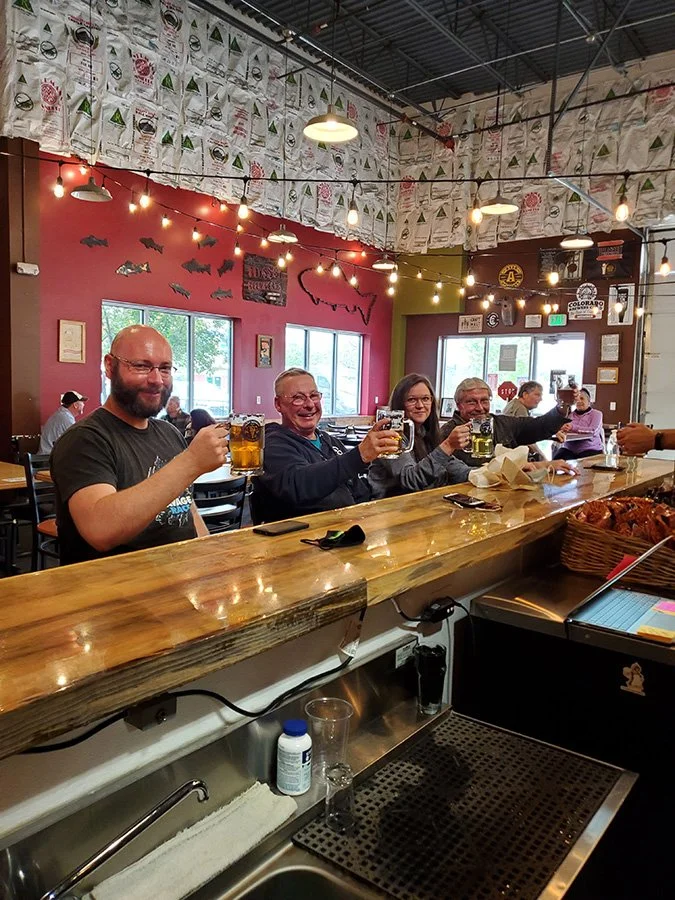 Four people sitting at a bar counter raising glasses of beer in a toast inside a lively restaurant with colorful decor, string lights, and large windows.