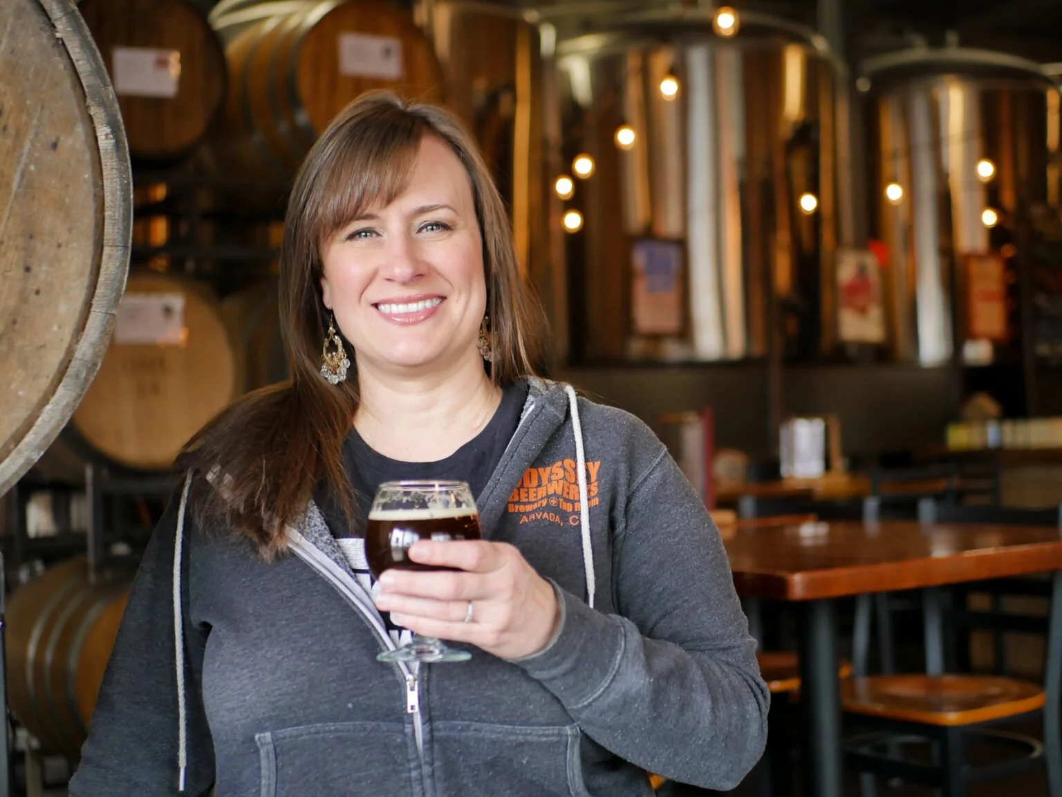 A smiling woman holding a glass of dark beer in a brewery or bar with wooden barrels and tables in the background.