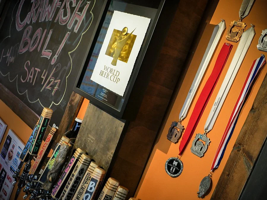 A wall with three medals hanging and a framed poster of a beer cup, with a chalkboard advertising a crawfish boil event scheduled for Saturday, April 24.