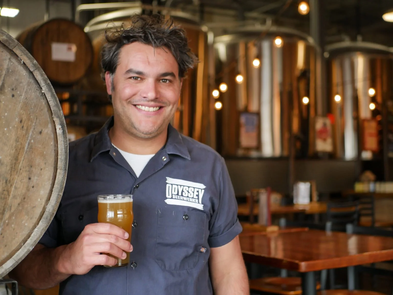 A smiling man in a gray work shirt with the logo "Odyssey Beerwerks" holding a glass of beer, standing inside a brewery with large wooden barrels and warm lighting in the background.