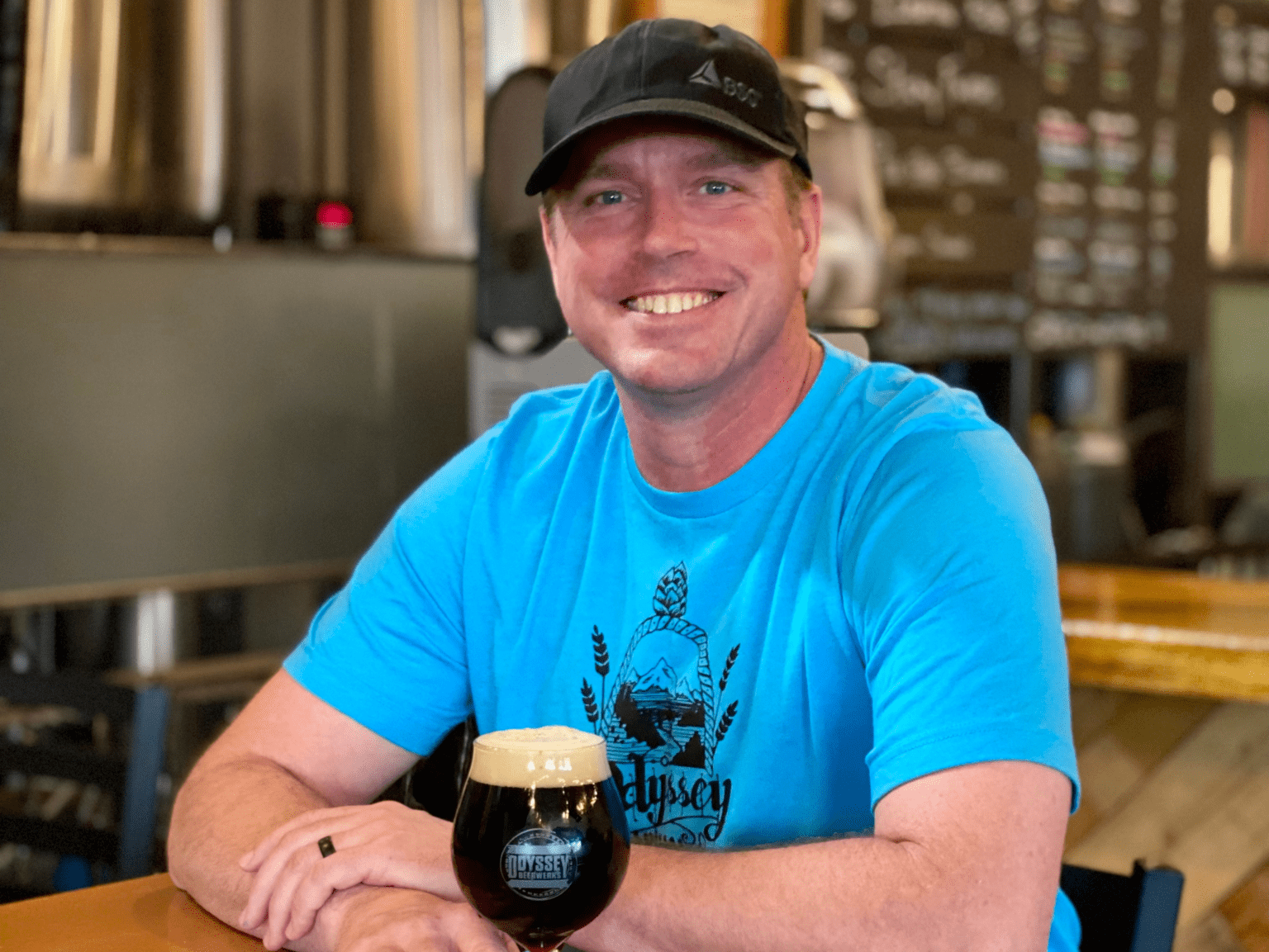A man smiling and sitting at a table in a brewery, wearing a black cap and a bright blue T-shirt, with a dark beer with a foamy head in front of him.