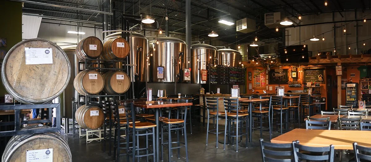 Interior of a brewery or restaurant with large stainless steel brewing tanks, wooden barrels, and a bar area with chairs and tables.