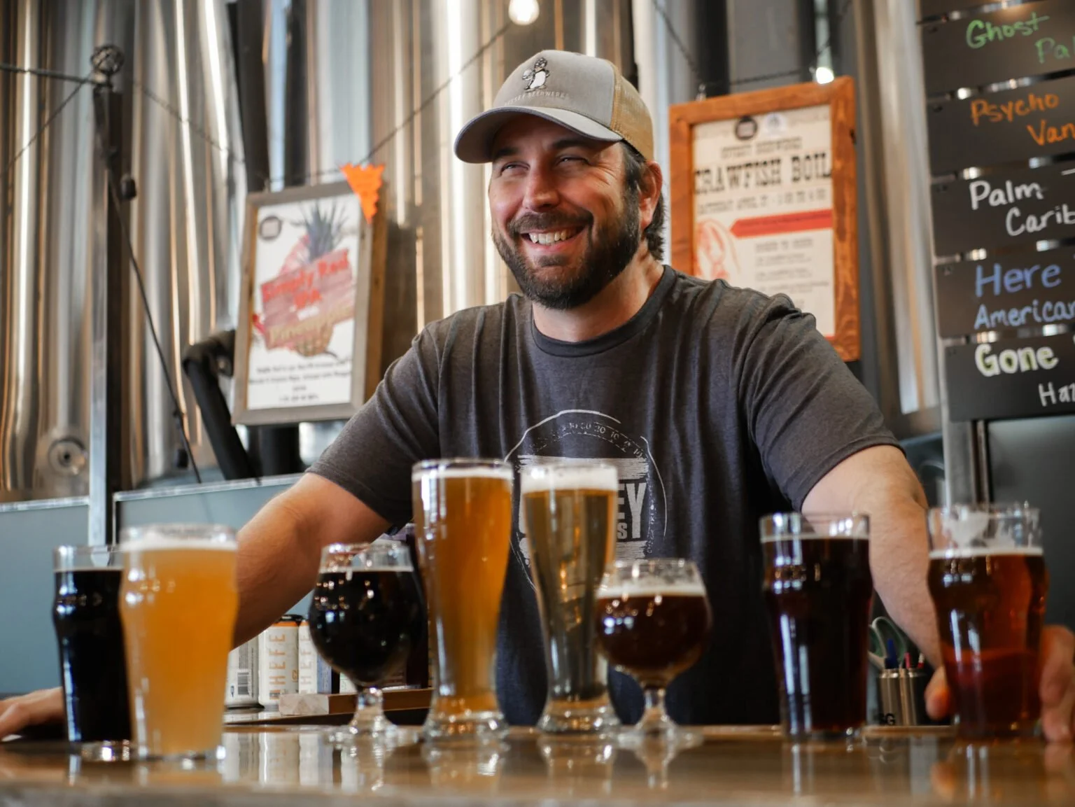 A smiling man with a beard wearing a gray cap and dark t-shirt standing behind a bar with various glasses of beer in front of him at a brewery or pub.