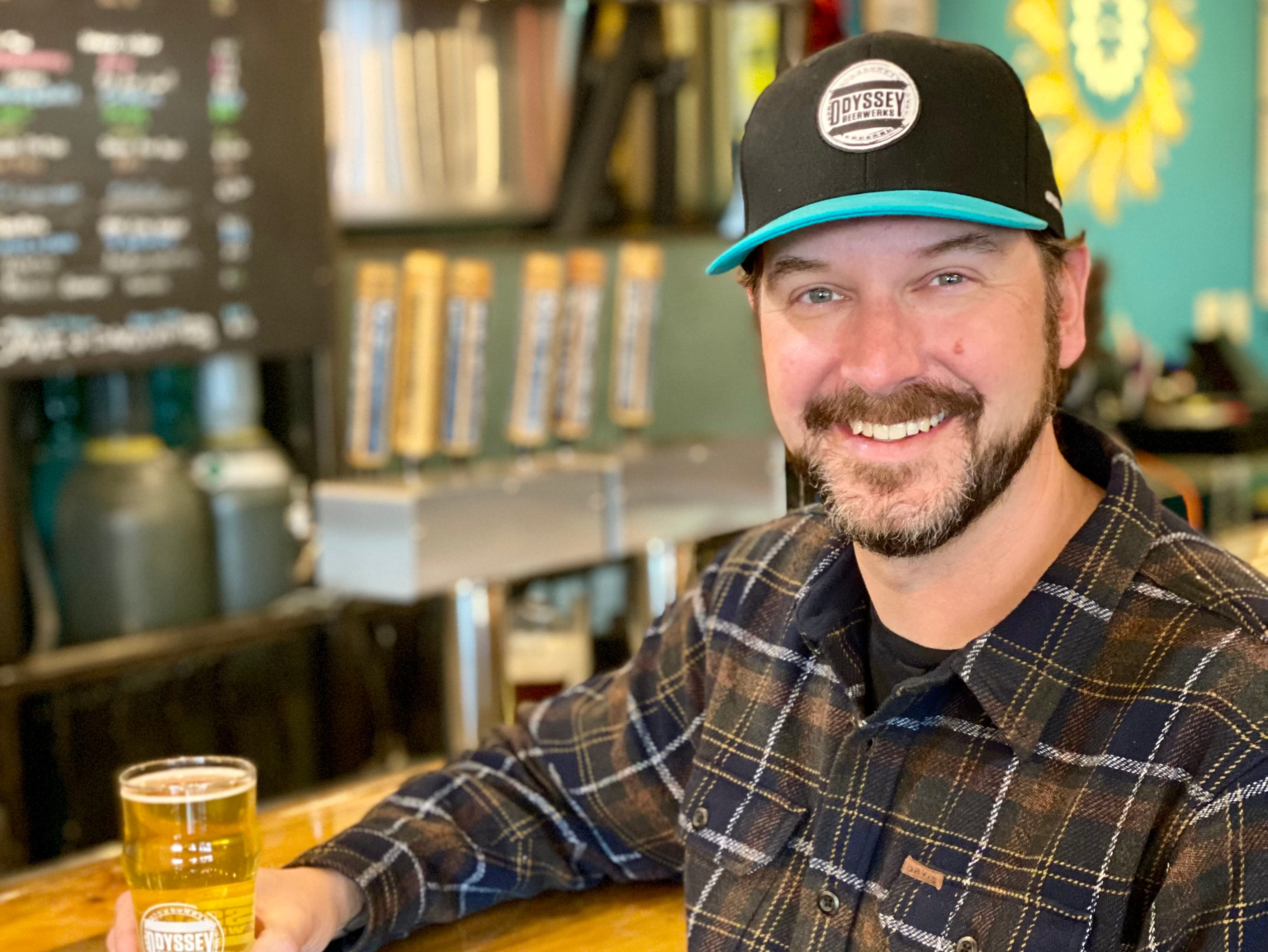 A smiling man with a beard and mustache sitting at a bar, holding a glass of beer, wearing a black cap with a logo, plaid shirt, in a brewery or bar setting.
