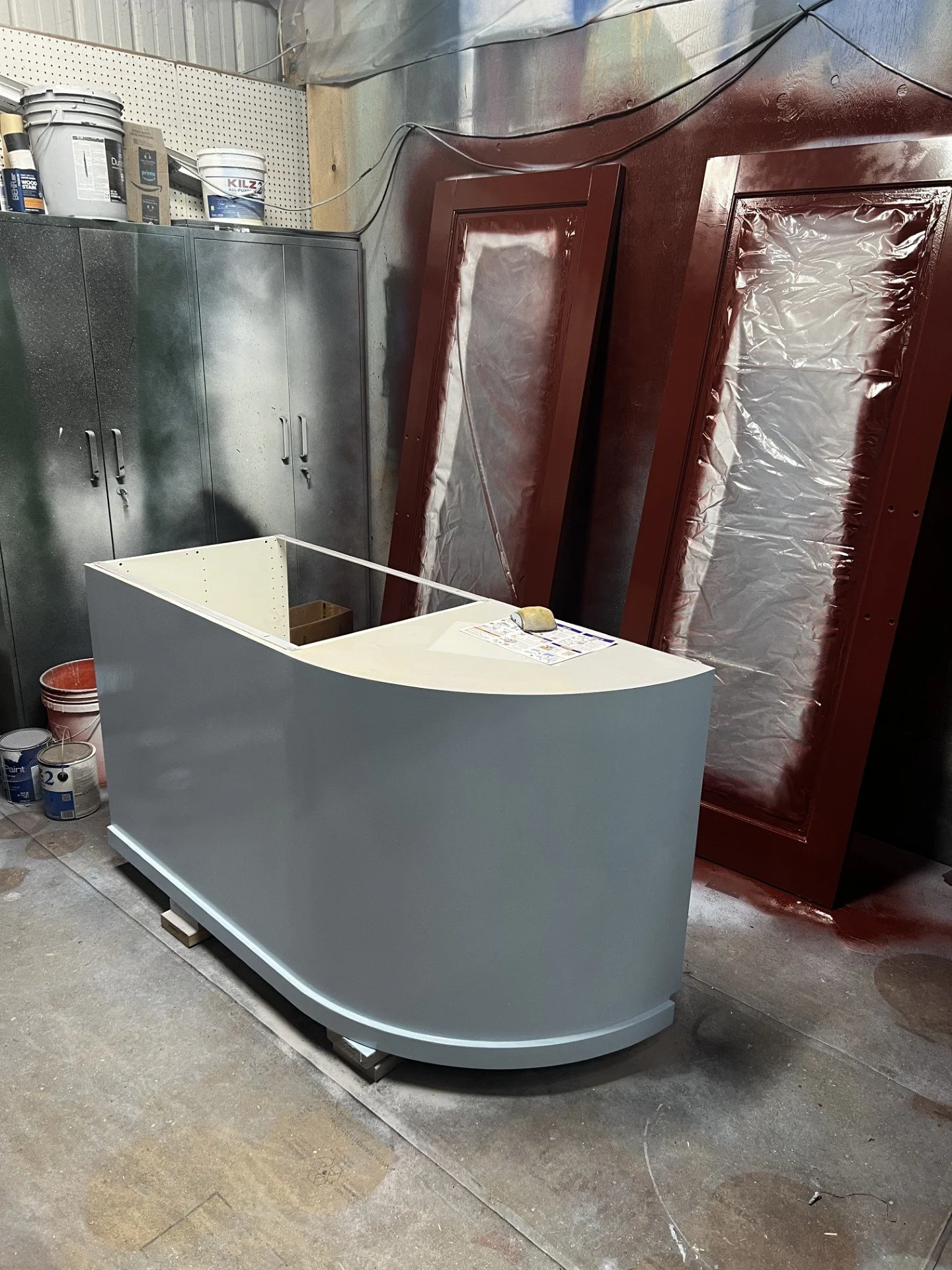 A curved white reception desk in a workshop with a paint roller on top, tall cabinets, and painting supplies, including stacked paint cans, against a wall with red and silver protective sheets.
