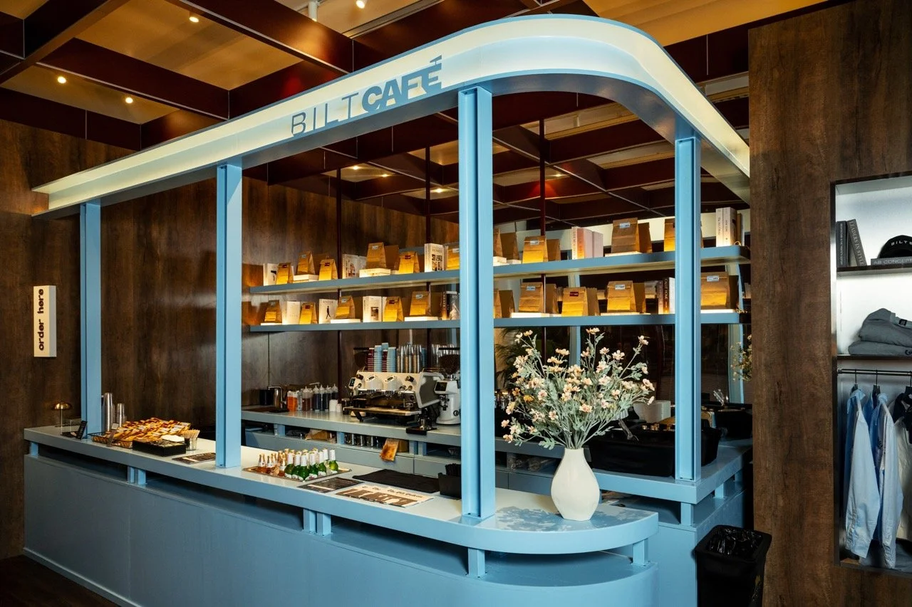 Blue café counter with a vase of flowers, coffee machine, and snacks inside BILT Café, with wooden wall and shelving behind.