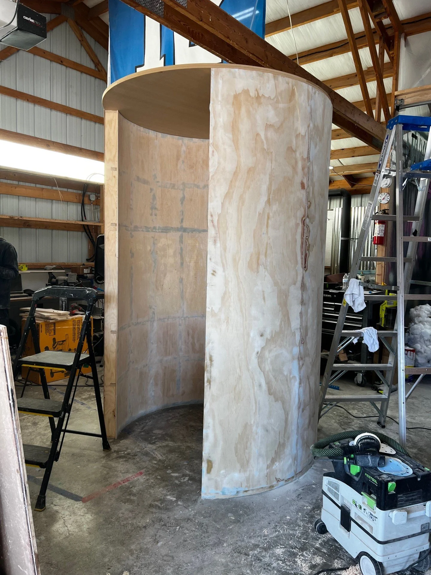 Construction of a curved wooden structure inside a workshop, with tools and ladders around.