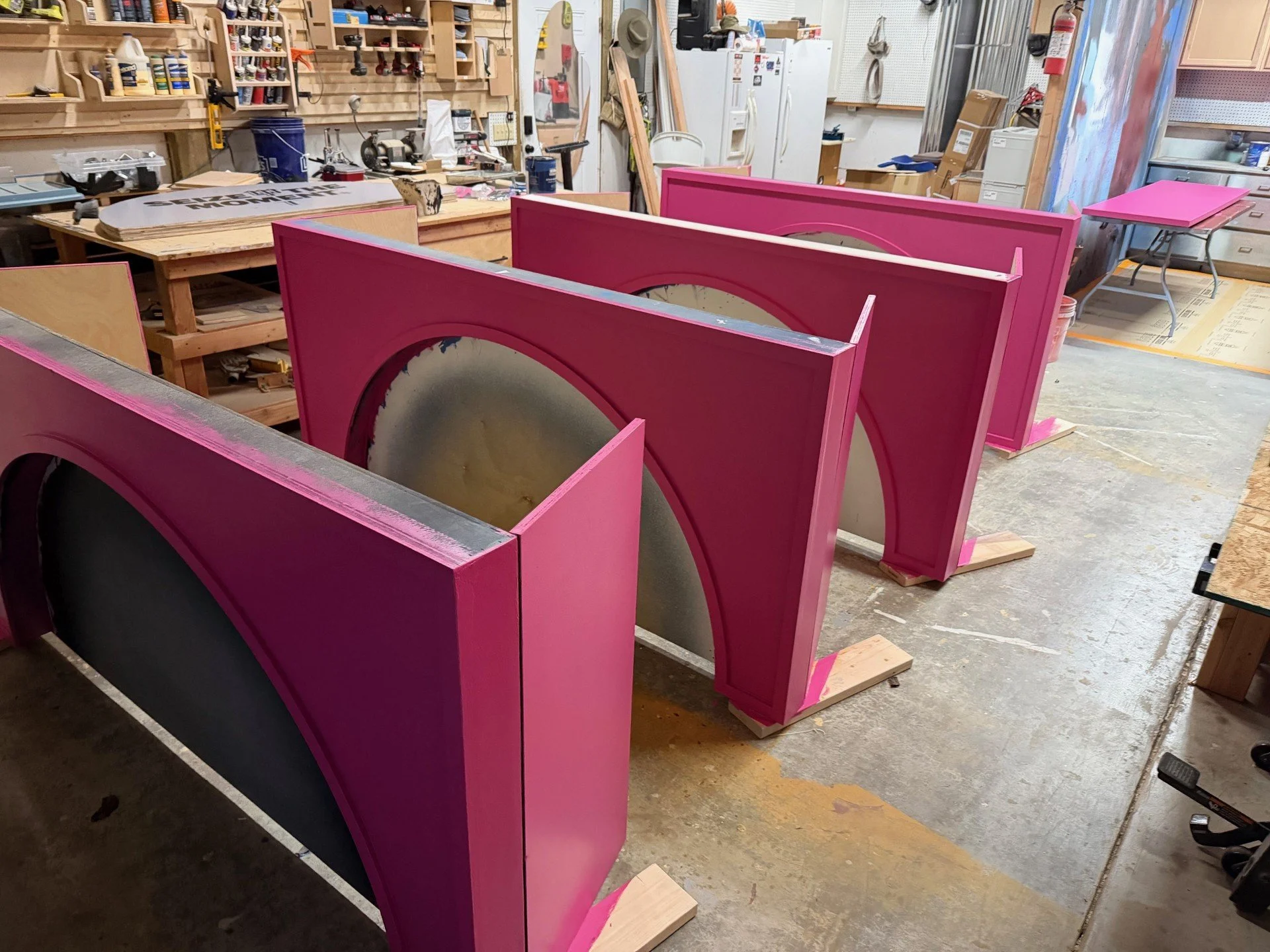 Workshop with pink-painted decorative panels with circular cutouts on wooden supports, surrounded by tools and woodworking equipment.