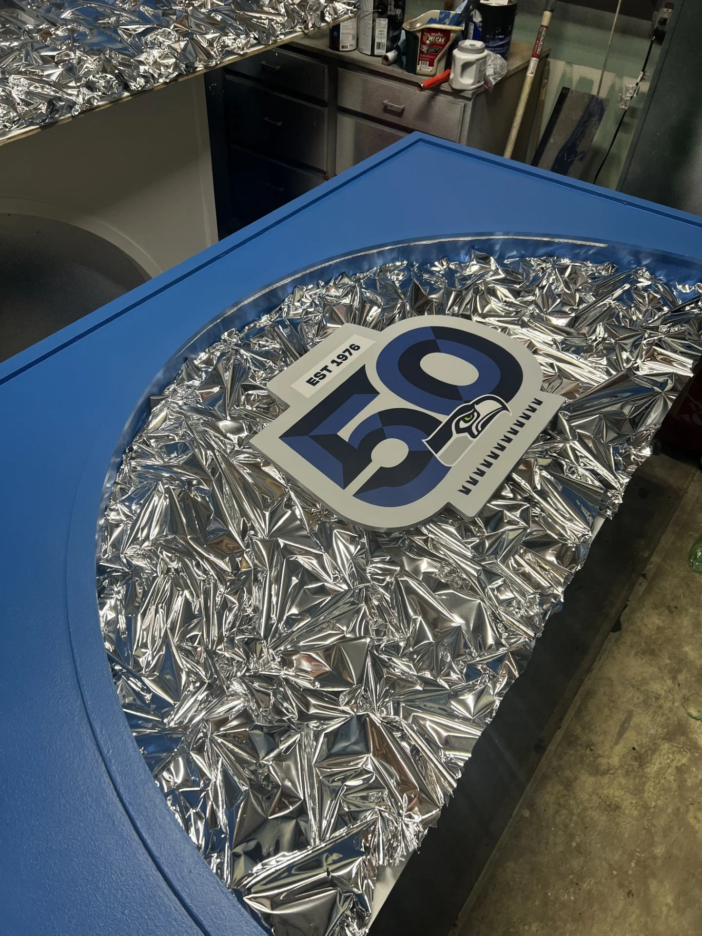 A blue ice chest with a lid covered in crinkled aluminum foil and a sticker that reads "Hawks" with a hawk logo. The surrounding area shows kitchen items and a laundry machine in the background.