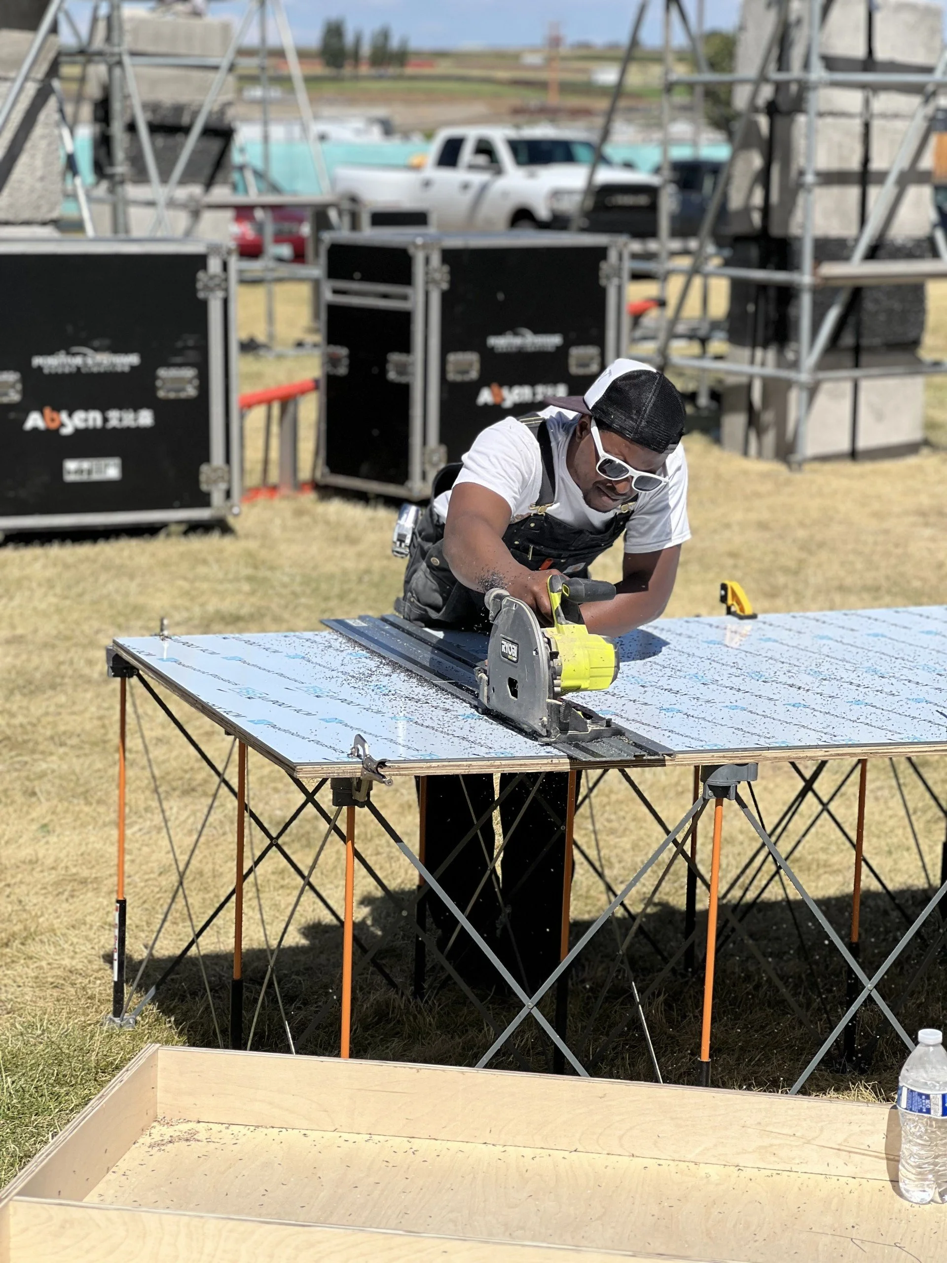 A man using a yellow saw to cut a piece of reflective material on a portable table outdoors, with equipment and trucks in the background.
