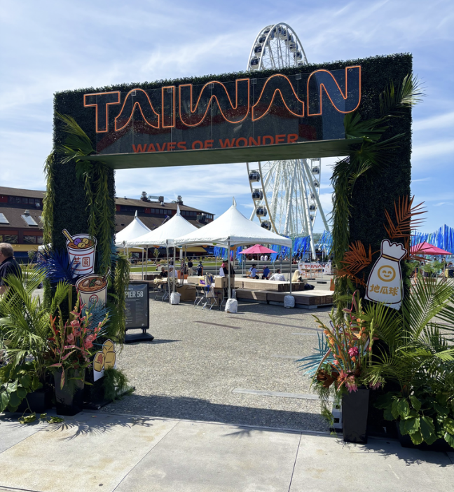 Entrance to Taiwan pavilion at a fair with a large Ferris wheel in the background, decorated with plants, tents, and booths.