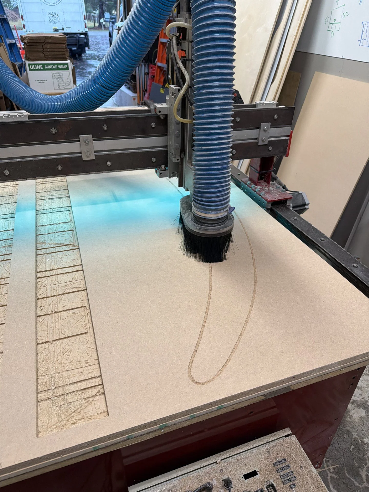 A CNC machine carving a curved design into a piece of light-colored wood or MDF in a workshop.