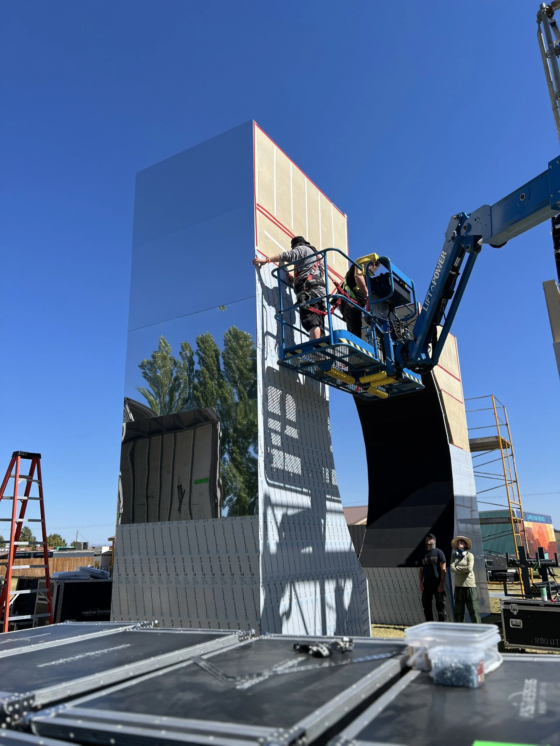 Workers install or work on a large, reflective, mirror-like structure with a black opening at the bottom, surrounded by equipment, ladders, and a clear blue sky.