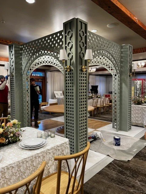 A decorative, freestanding partition with intricate lattice and arch designs inside a restaurant or event space. It has two lamps attached and is surrounded by tables, chairs, and floral arrangements.