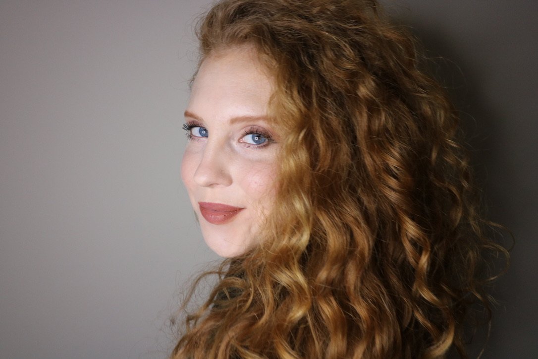 A woman with long, curly red hair and blue eyes smiling at the camera against a plain background.