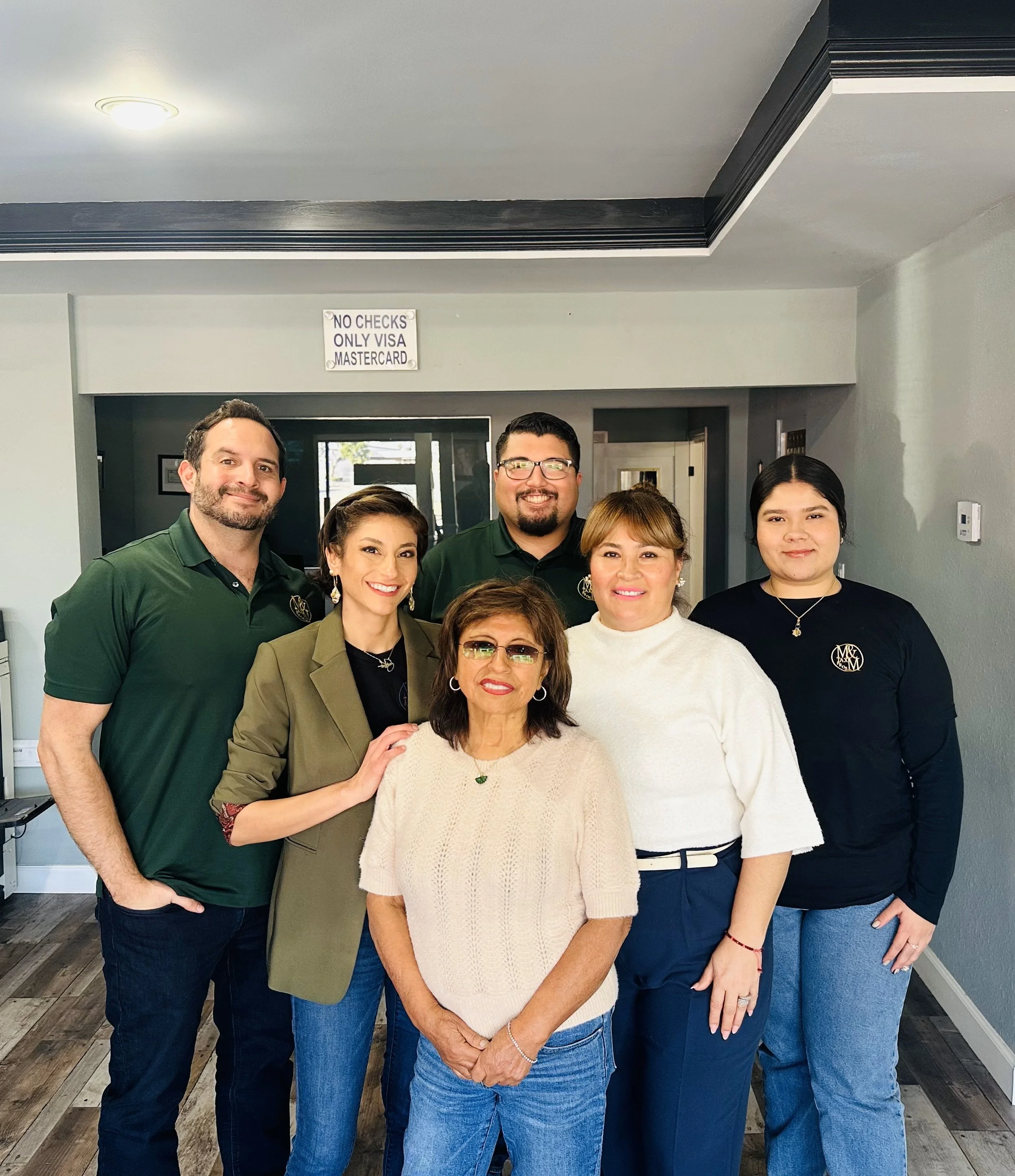 Six people standing inside a building, smiling at the camera, with a sign above the doorway that reads 'No Checks, Only Visa MasterCard'.