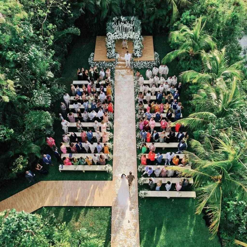 An outdoor wedding ceremony with guests seated on white benches on either side of a flower-filled aisle, surrounded by lush green trees and palms, with the bride and groom walking down the aisle towards the altar.