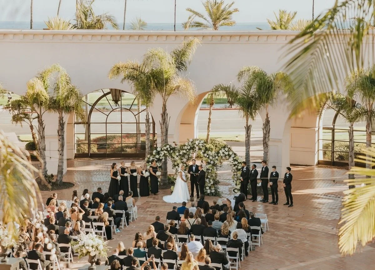 A wedding ceremony taking place outdoors under an arch with white flowers, with the bride and groom standing in front of an officiant. Bridesmaids and groomsmen stand beside them, opposite rows of seated guests, with tall palm trees and a building with large windows in the background.