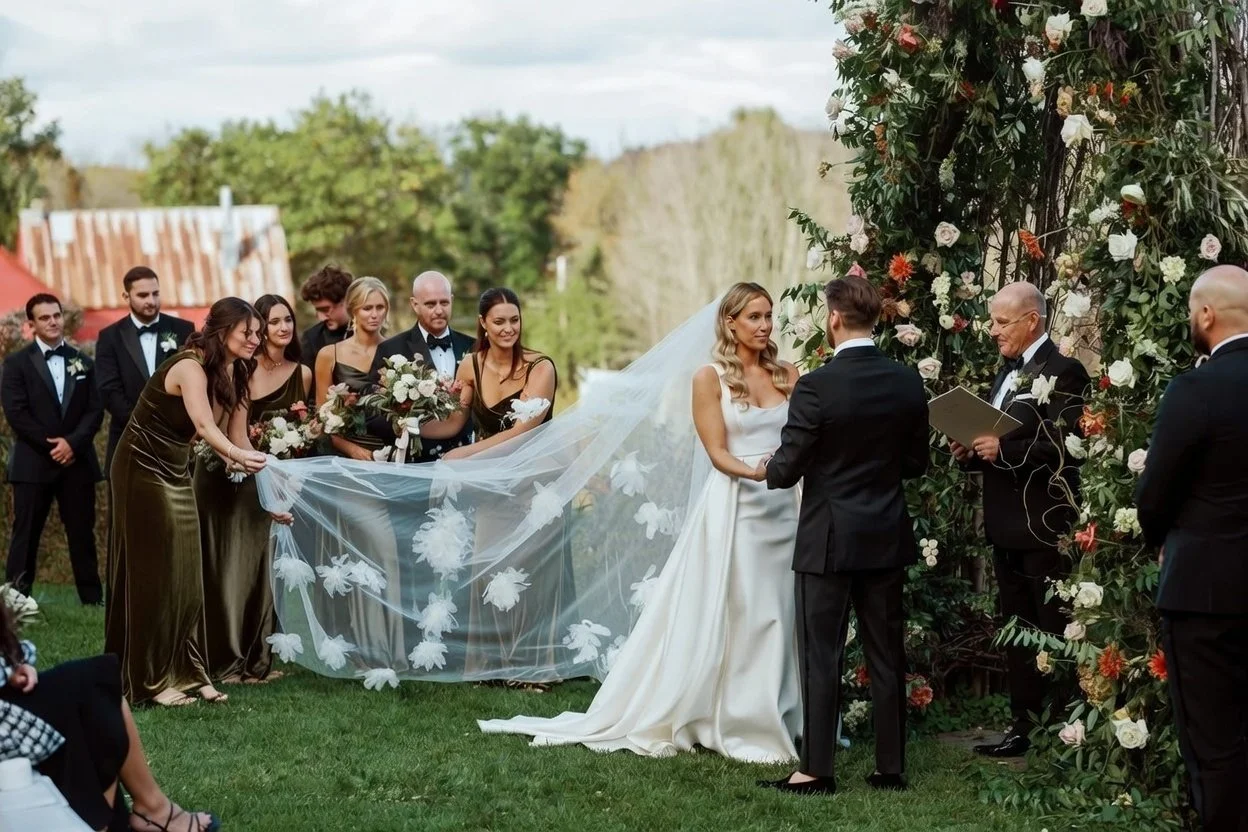 A wedding ceremony outdoors with the bride and groom exchanging vows in front of an arch of flowers. The bride is wearing a white wedding dress and veil, while the groom is in a black suit. Bridesmaids in gold dresses hold a long floral veil, and groomsmen in black suits stand nearby. The scene is set in a lush green garden with trees in the background.