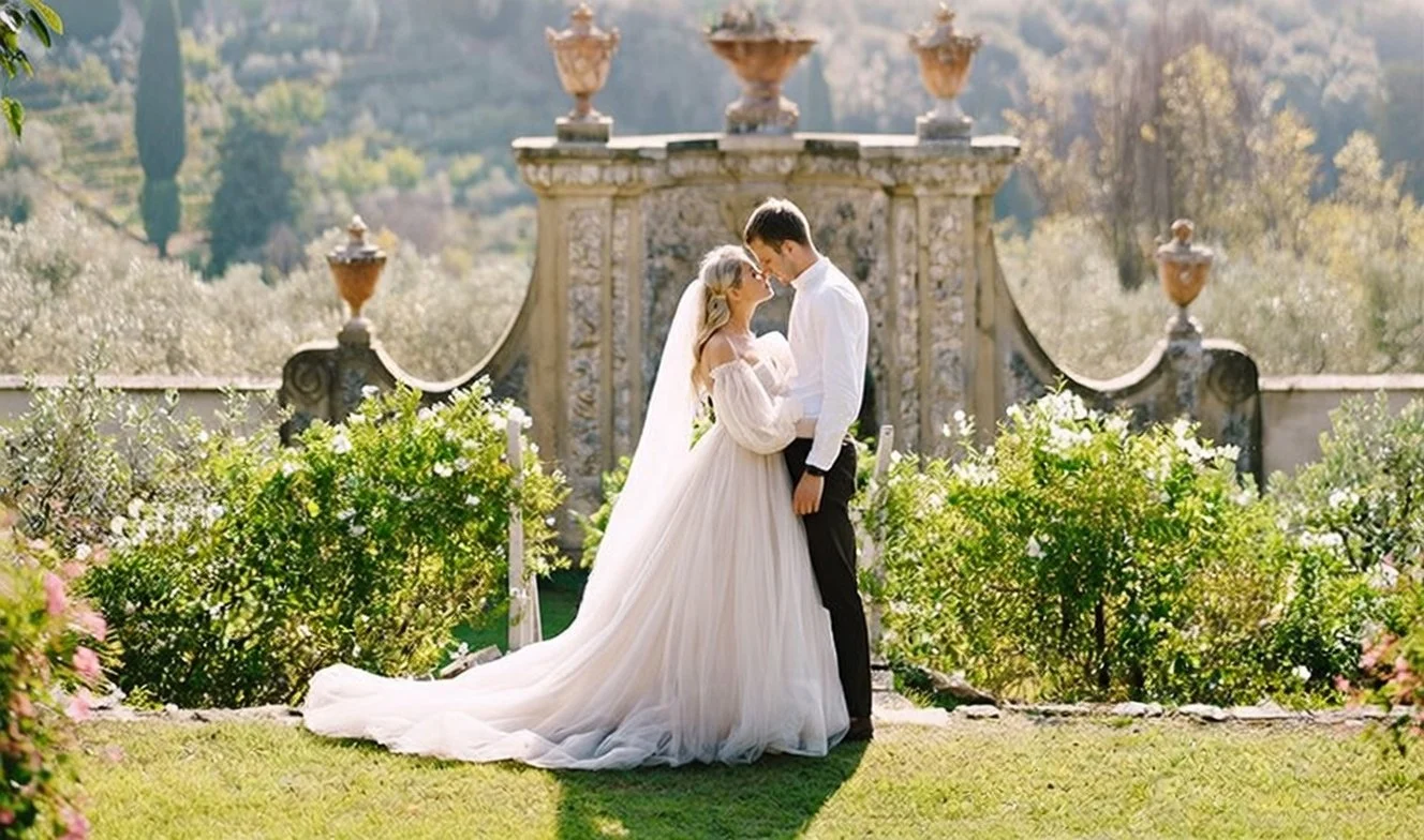 A bride and groom standing close together, facing each other with foreheads touching, outdoors in a garden setting with ornate stone architecture and lush greenery in the background.