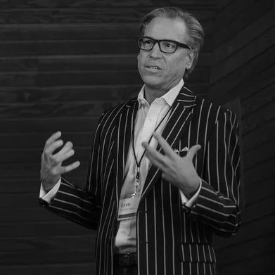 A man in a striped suit and glasses speaking or presenting in front of a dark wooden wall.