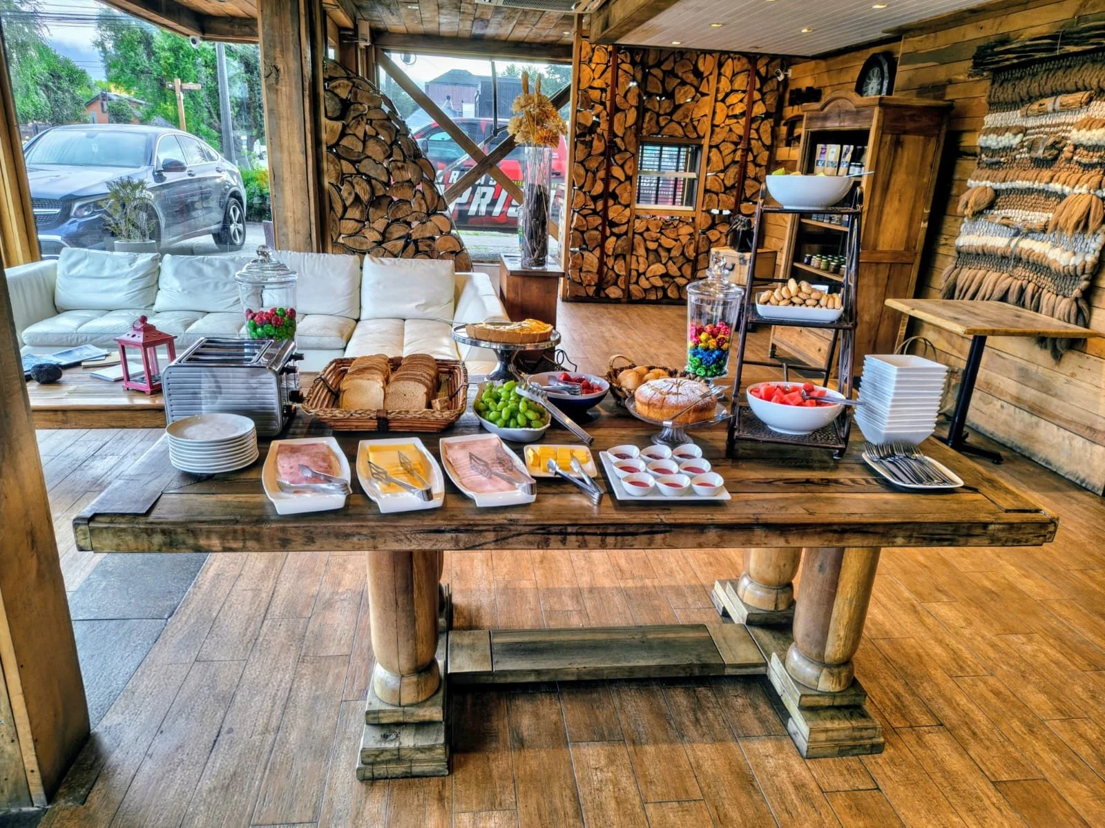 Una mesa de desayuno con varias rebanadas de pan, huevos, fruta, tocino, y diversos cereales y dulces, en un ambiente rústico con madera.