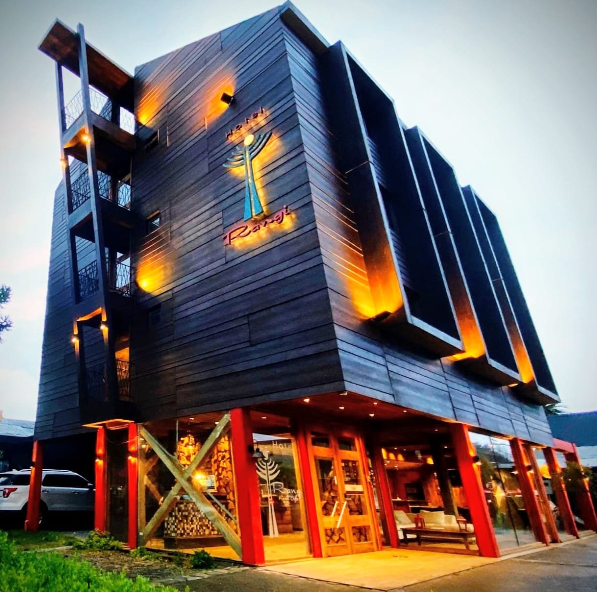 Edificio moderno de varios pisos con fachada de madera oscura y ventanas grandes, con luminarias cálidas. En la parte inferior, un restaurante con estructura de madera y columnas rojas, con puertas de cristal y decoraciones interiores visibles.