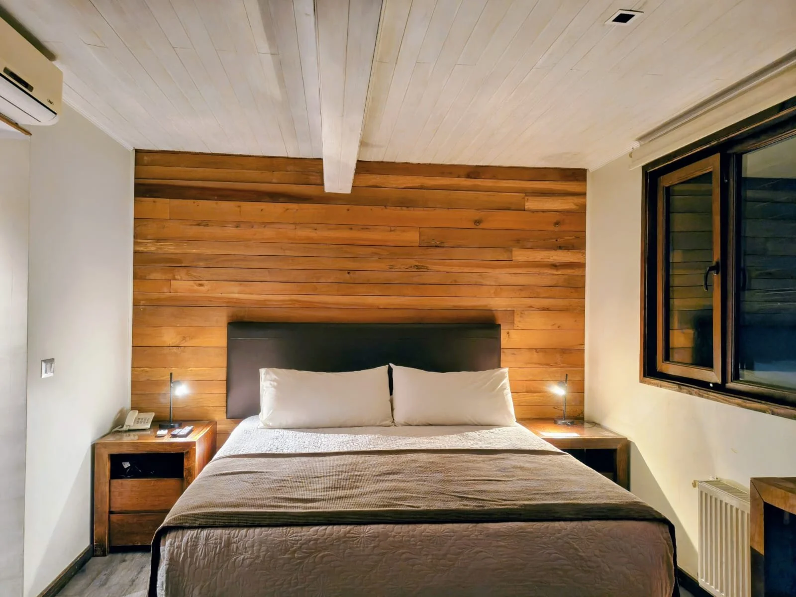 Habitación con cama doble, cabecera de color negro, paredes de madera, dos lámparas de noche, ventana con marco de madera, aire acondicionado