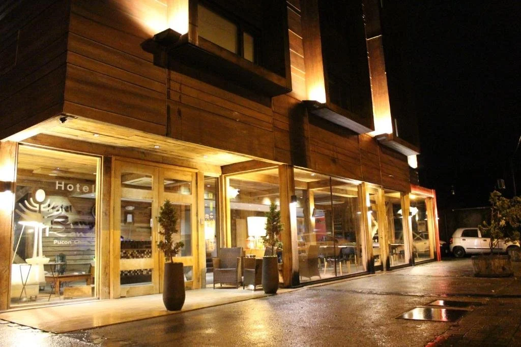 Fachada de un hotel con ventanas grandes y puertas de madera, interior iluminado, plantas decorativas en macetas negras y coches estacionados afuera en la noche.