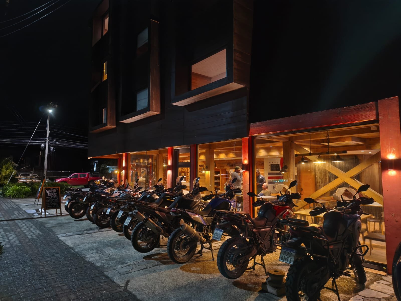 Fachada de un restaurante o cafetería con muchas motos estacionadas afuera, iluminada con luces cálidas, y un ambiente acogedor en la noche.