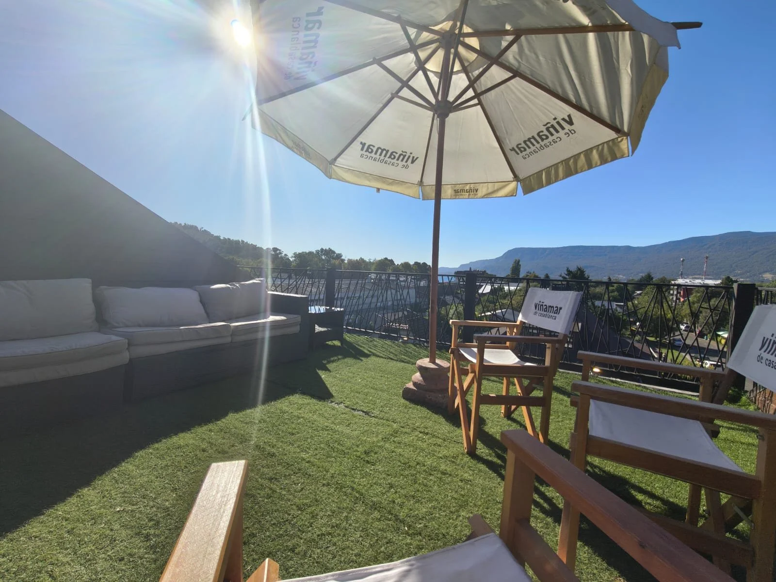 Terraza al aire libre con sillones de madera y respaldo de lona, una sombrilla grande con logo de Vinamar, vista de montañas y cielo despejado, ambiente soleado y relajante.