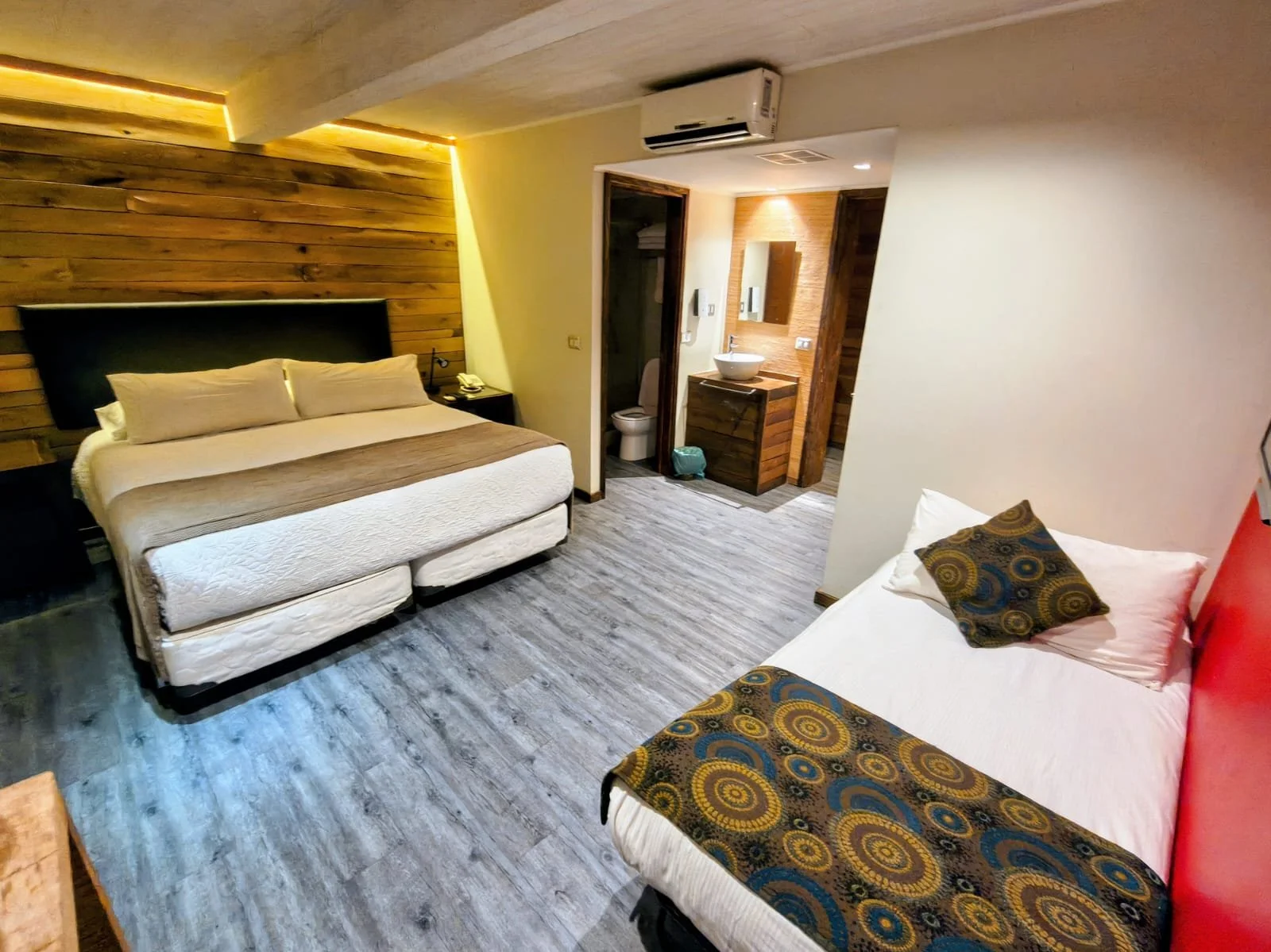 Habitación de hotel con dos camas, un baño visible y paredes de madera, decorada con estilo moderno y cálido.